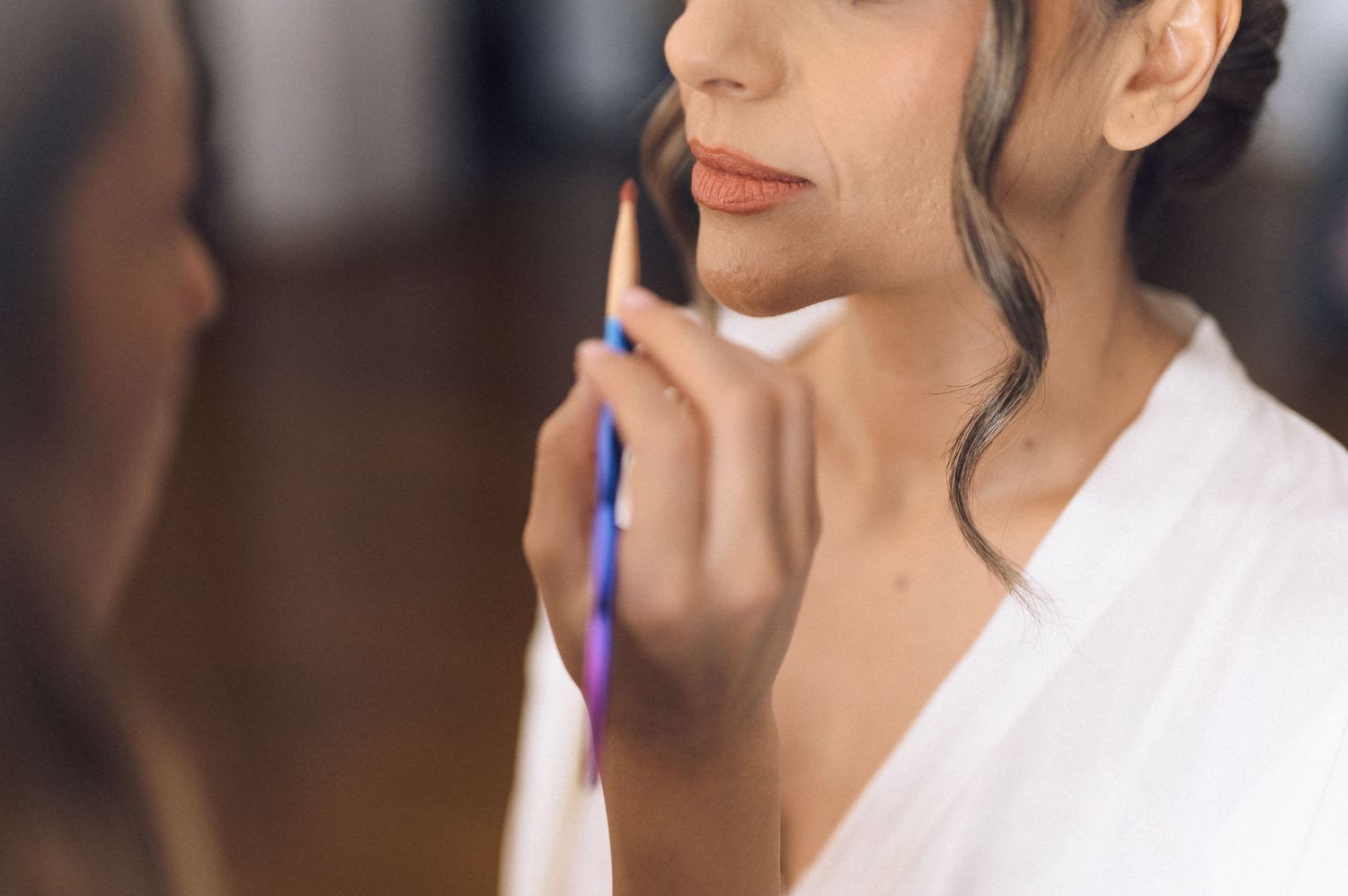 A bride in a white robe applies makeup with a brush while looking in a mirror during wedding preparations.