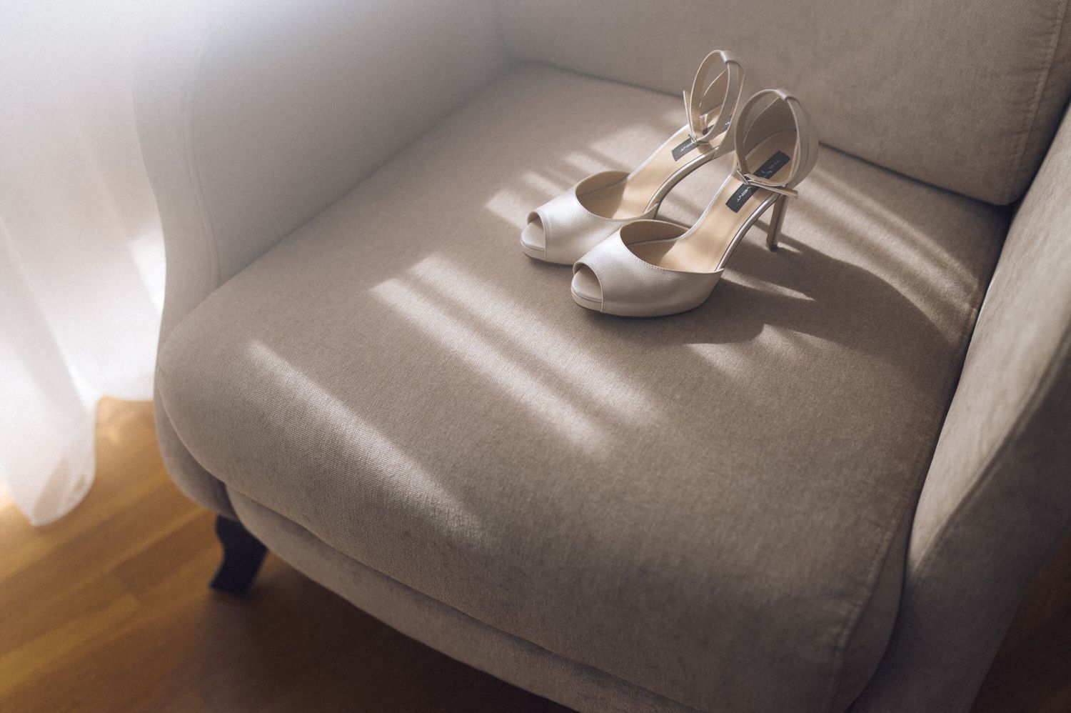 Elegant white strappy high heel sandals resting on a gray upholstered chair in soft natural light.