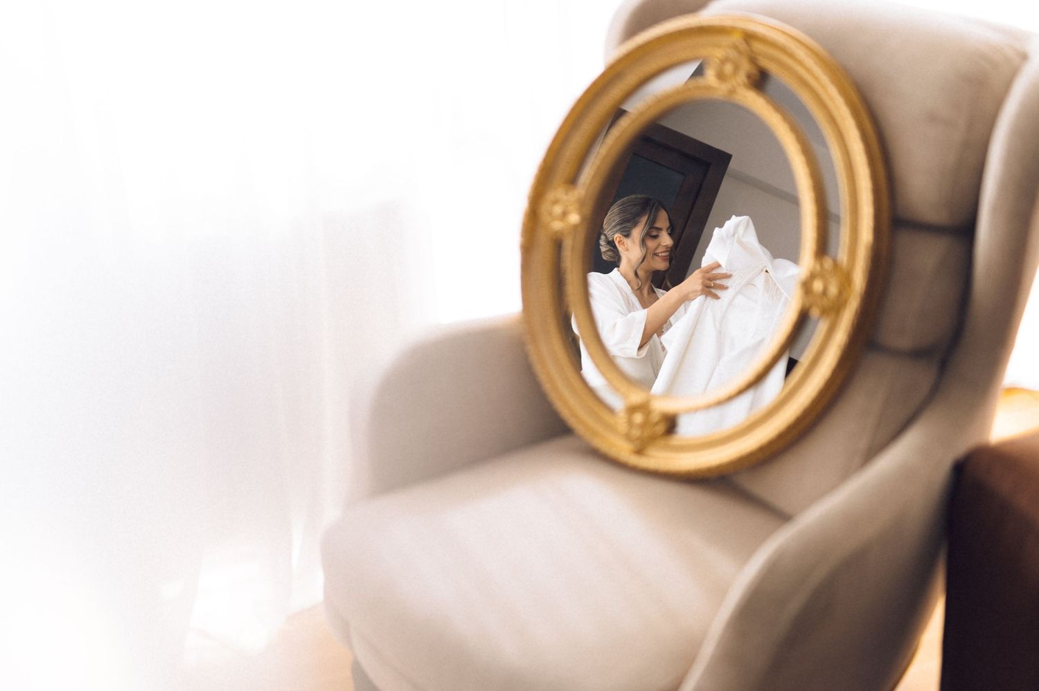 A woman in a white robe holds a wedding dress while viewing her reflection in an ornate gold oval mirror on a chair.