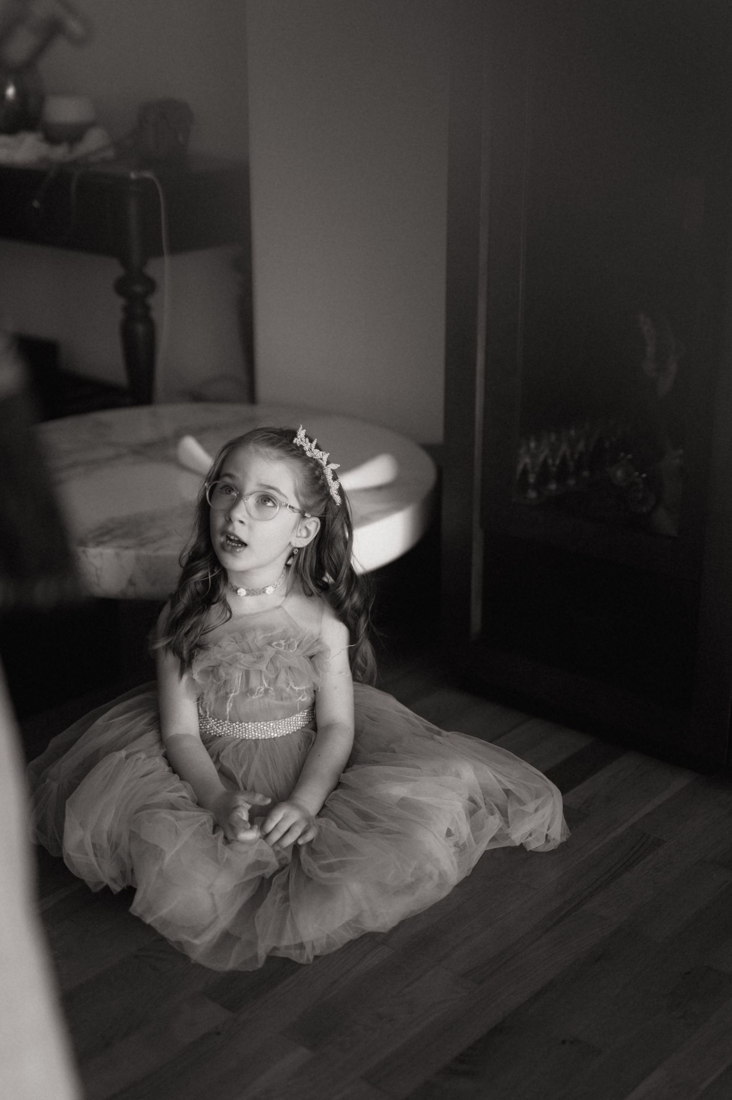 Black and white photo of a young girl in a tulle dress and tiara sitting on a hardwood floor looking up.
