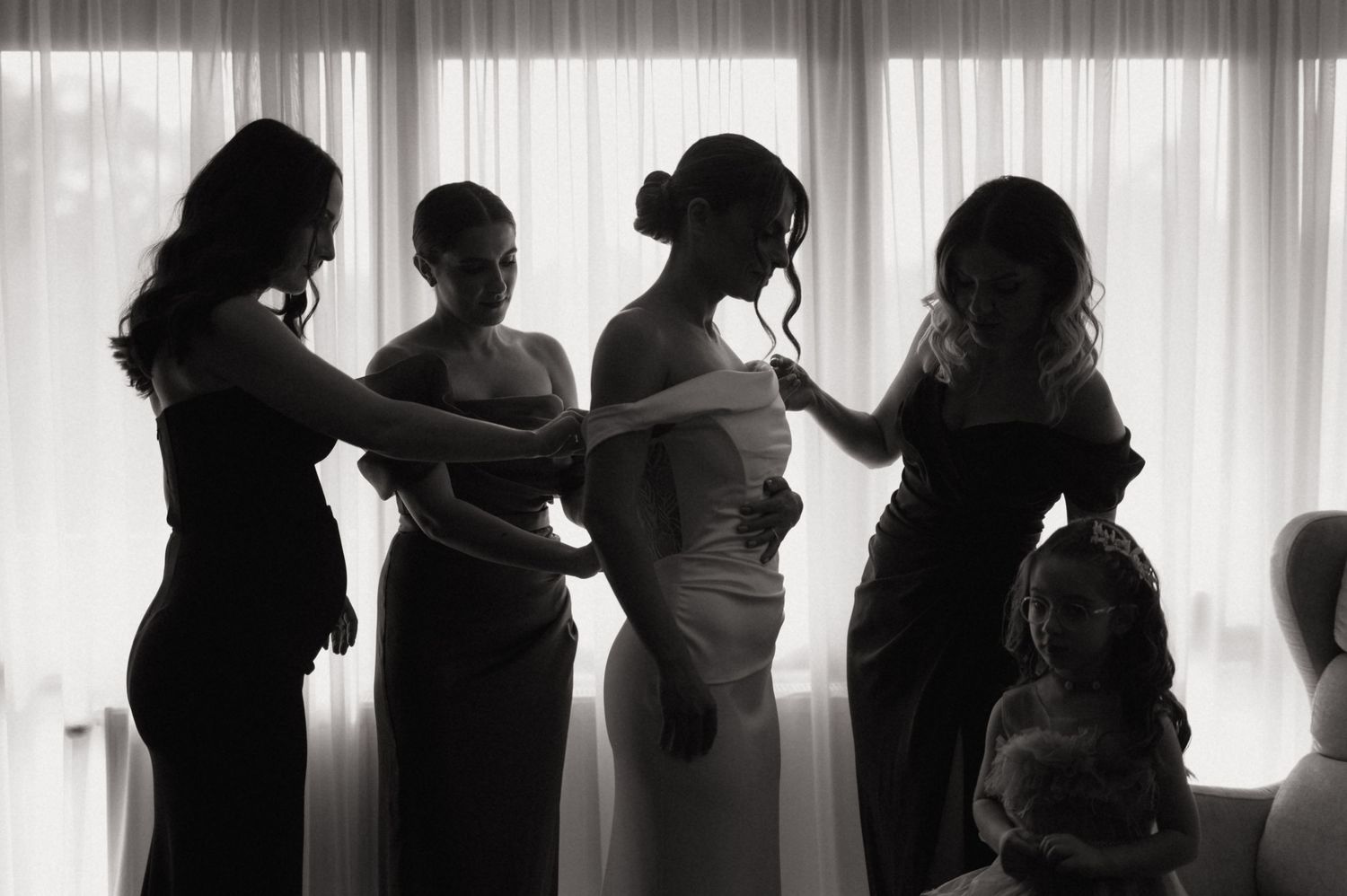 Black and white photo of bridesmaids helping bride into wedding dress by window.