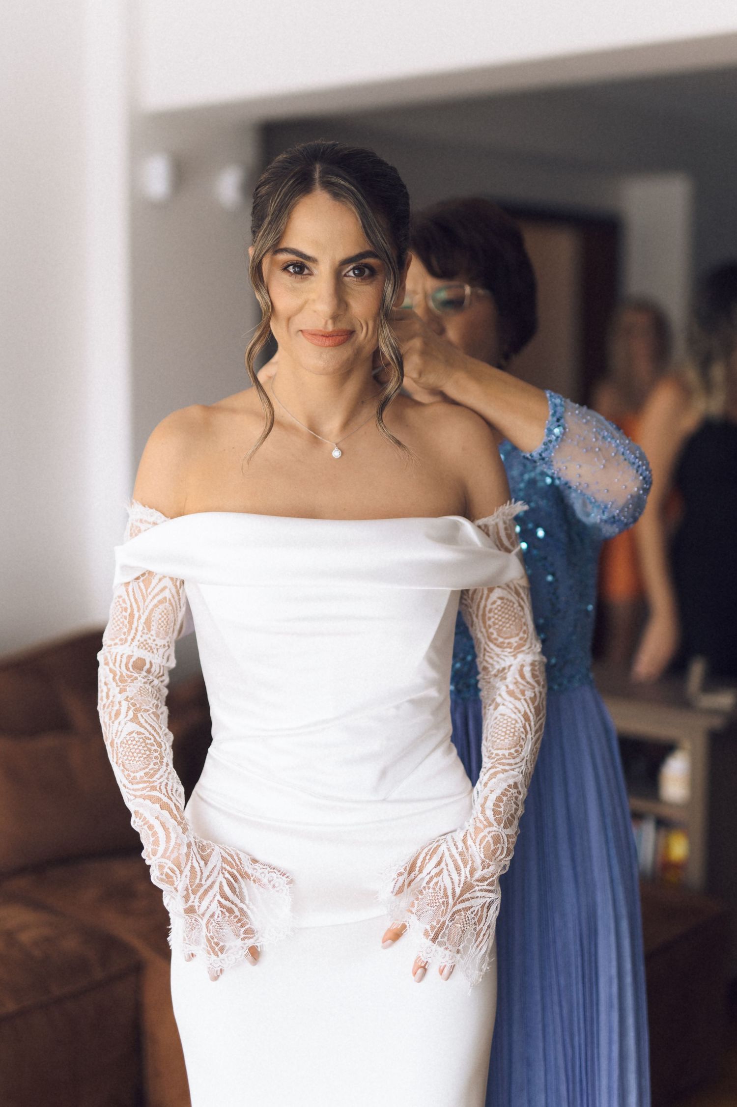 Bride in off-shoulder white lace wedding gown being helped by bridesmaid in blue dress during bridal preparations.