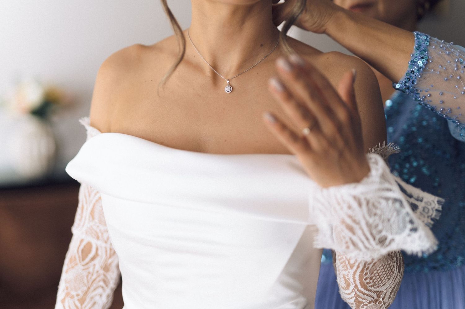 Close-up of bride in white off-shoulder lace wedding dress with delicate pearl necklace being assisted by bridesmaid.