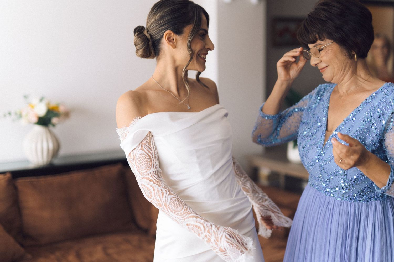 Bride in white lace gown stands with emotional mother in blue dress during wedding preparation.