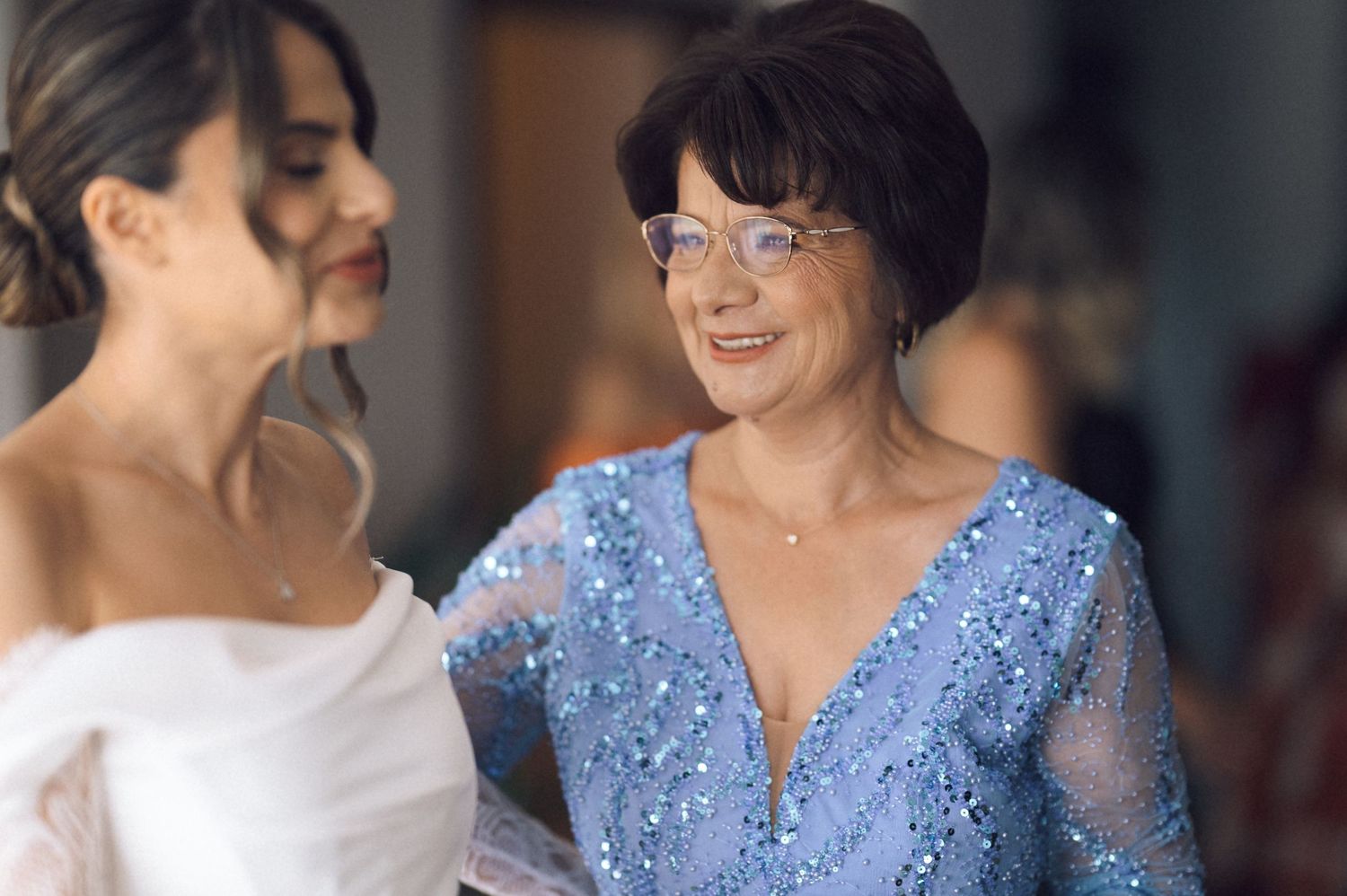Bride in white gown smiles with older woman in blue sequin dress during wedding preparations.
