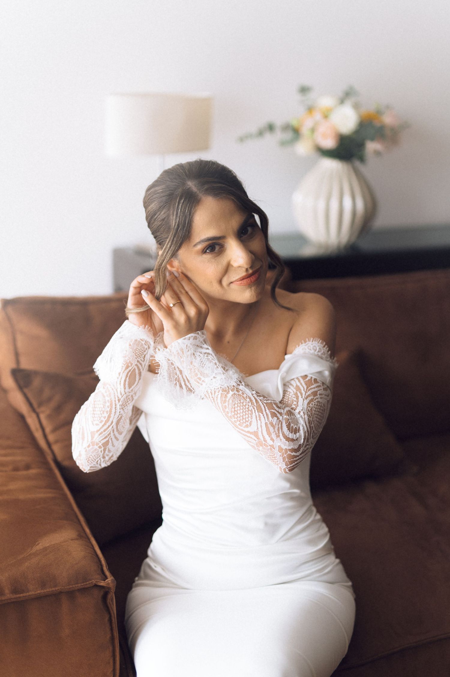 Bride in white off-shoulder lace wedding gown sitting on brown leather sofa, touching her ear elegantly.