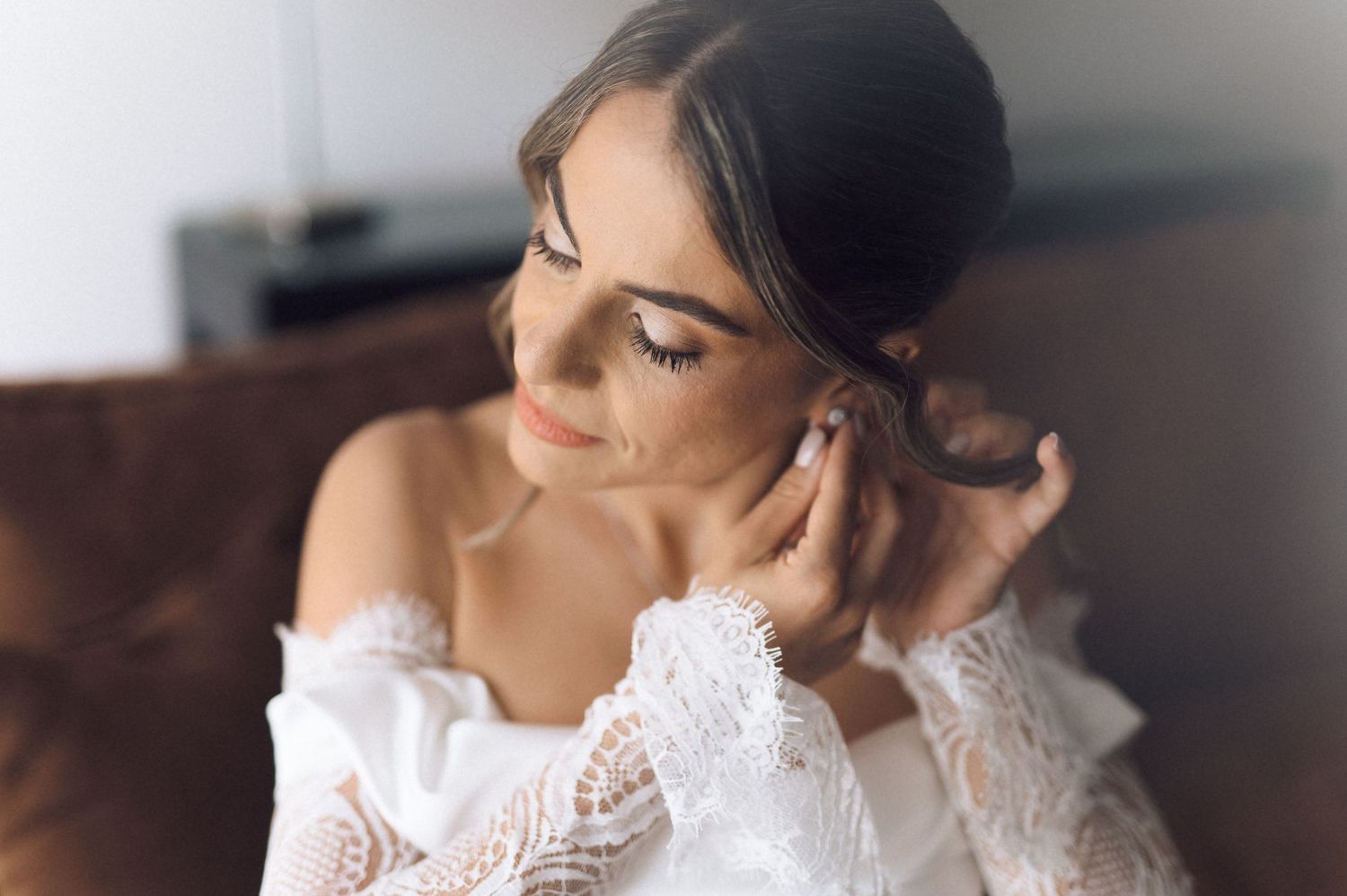 Bride in lace off-shoulder gown adjusting pearl earring, soft natural light, elegant bridal portrait.