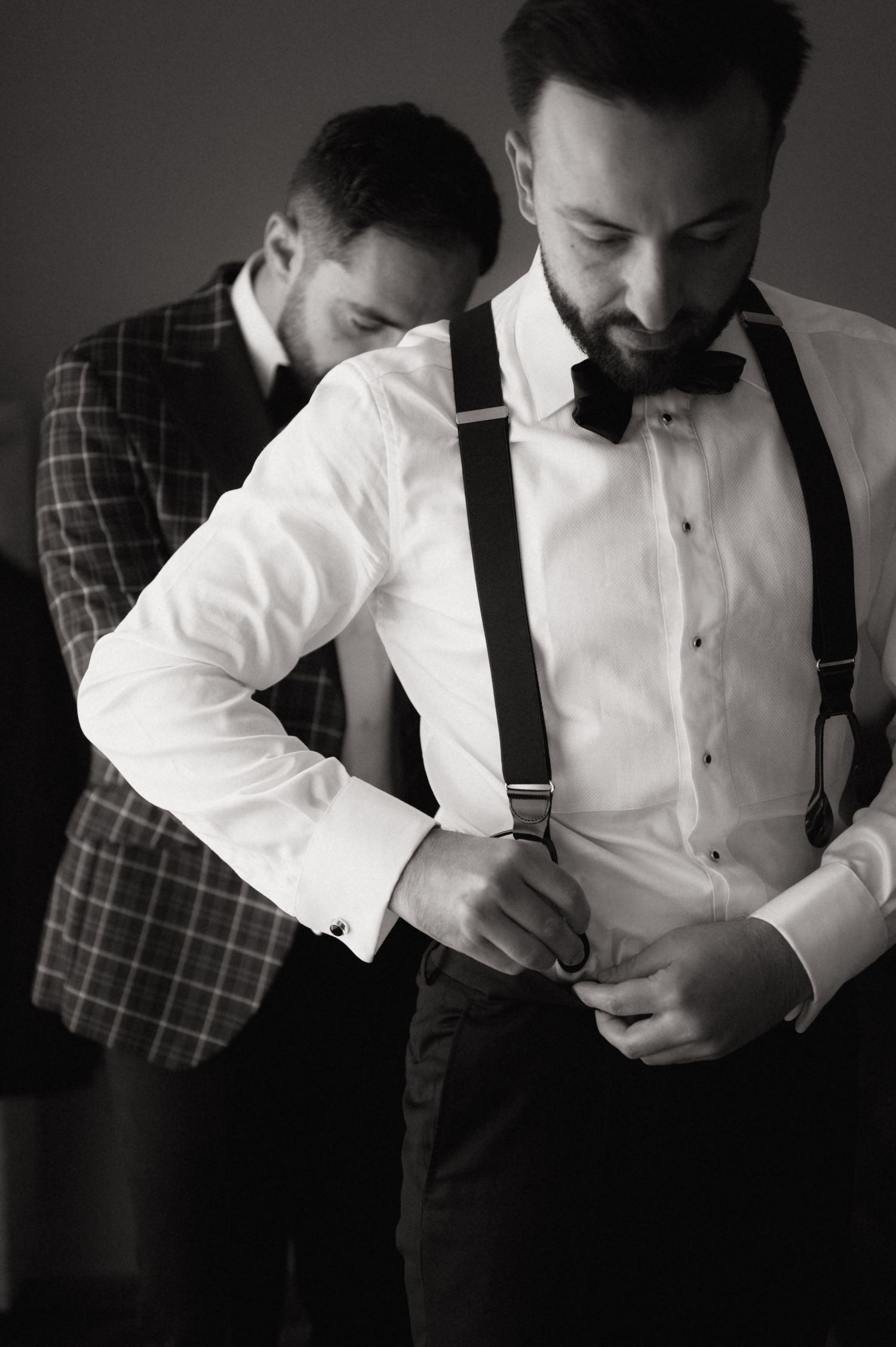 Groom in white shirt, suspenders, and bow tie adjusting buttons while best man in plaid suit assists during wedding preparations.
