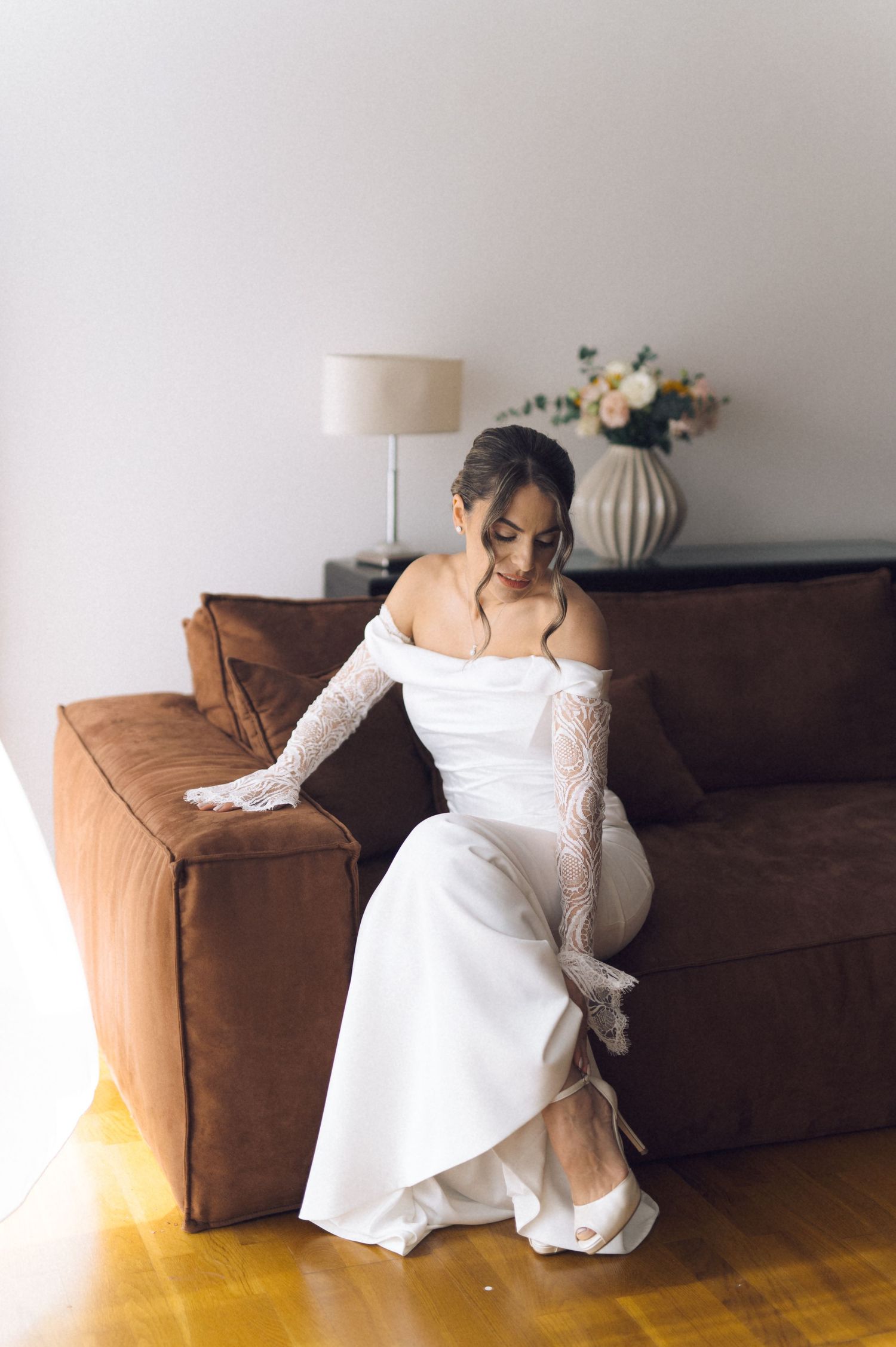 Bride in white off-shoulder gown with lace gloves sitting gracefully on a brown velvet sofa.