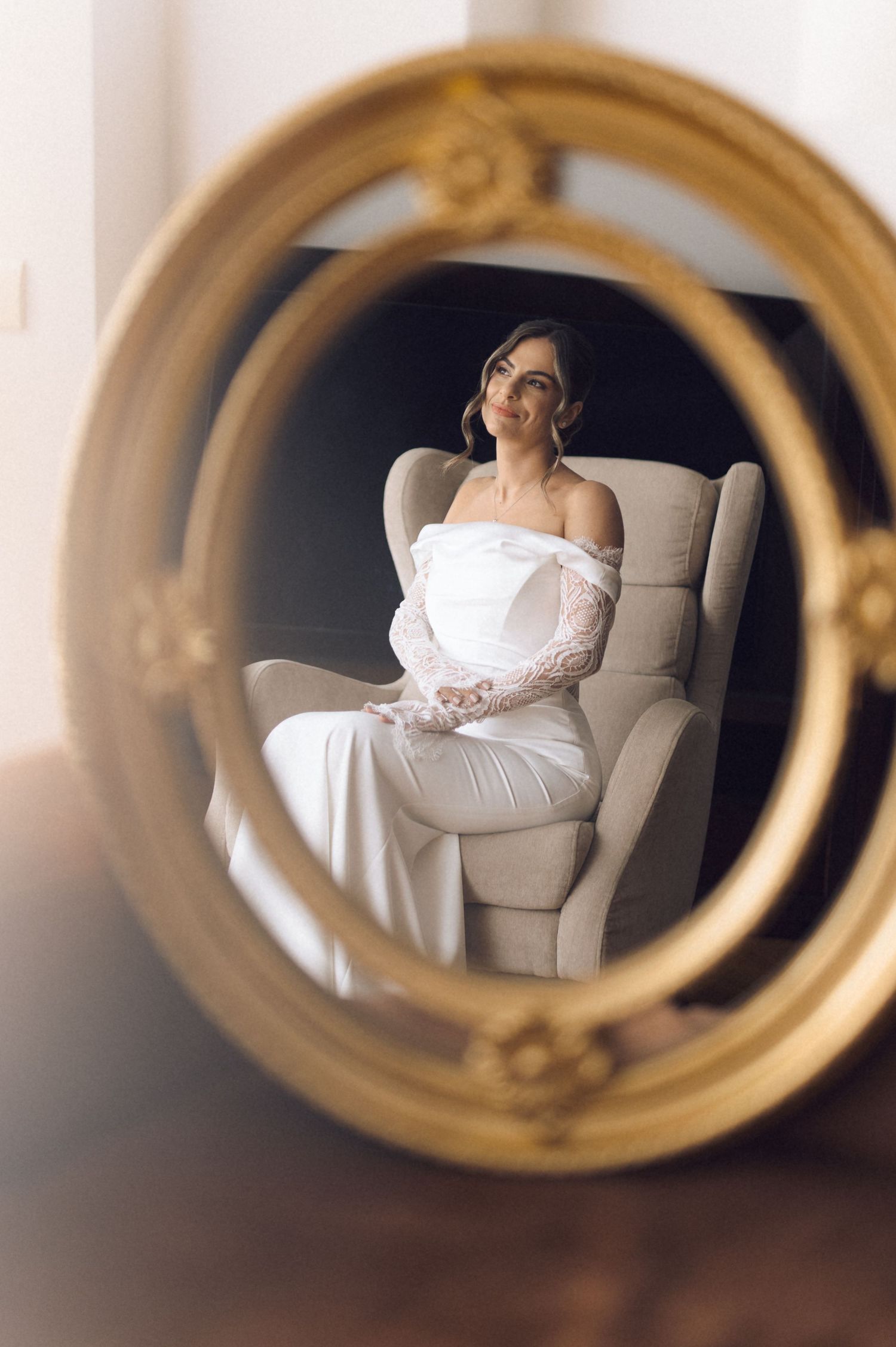 Bride in white gown and lace gloves seated in armchair, reflected in ornate gold oval mirror.