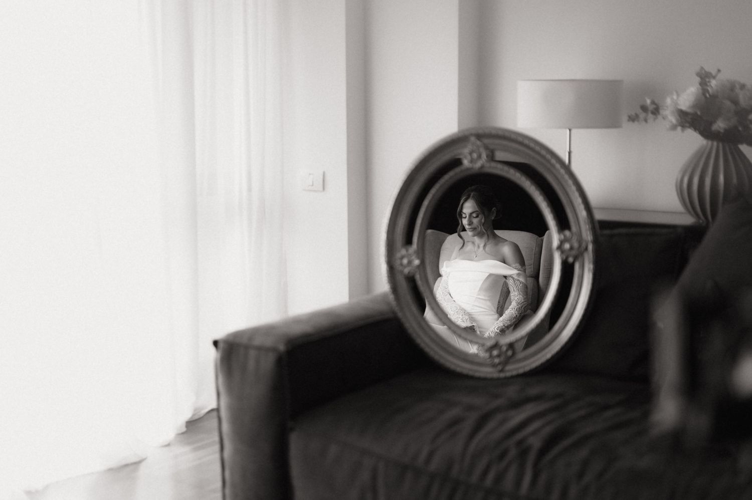 Black and white photo of a pregnant woman reflected in an ornate oval mirror on a sofa.