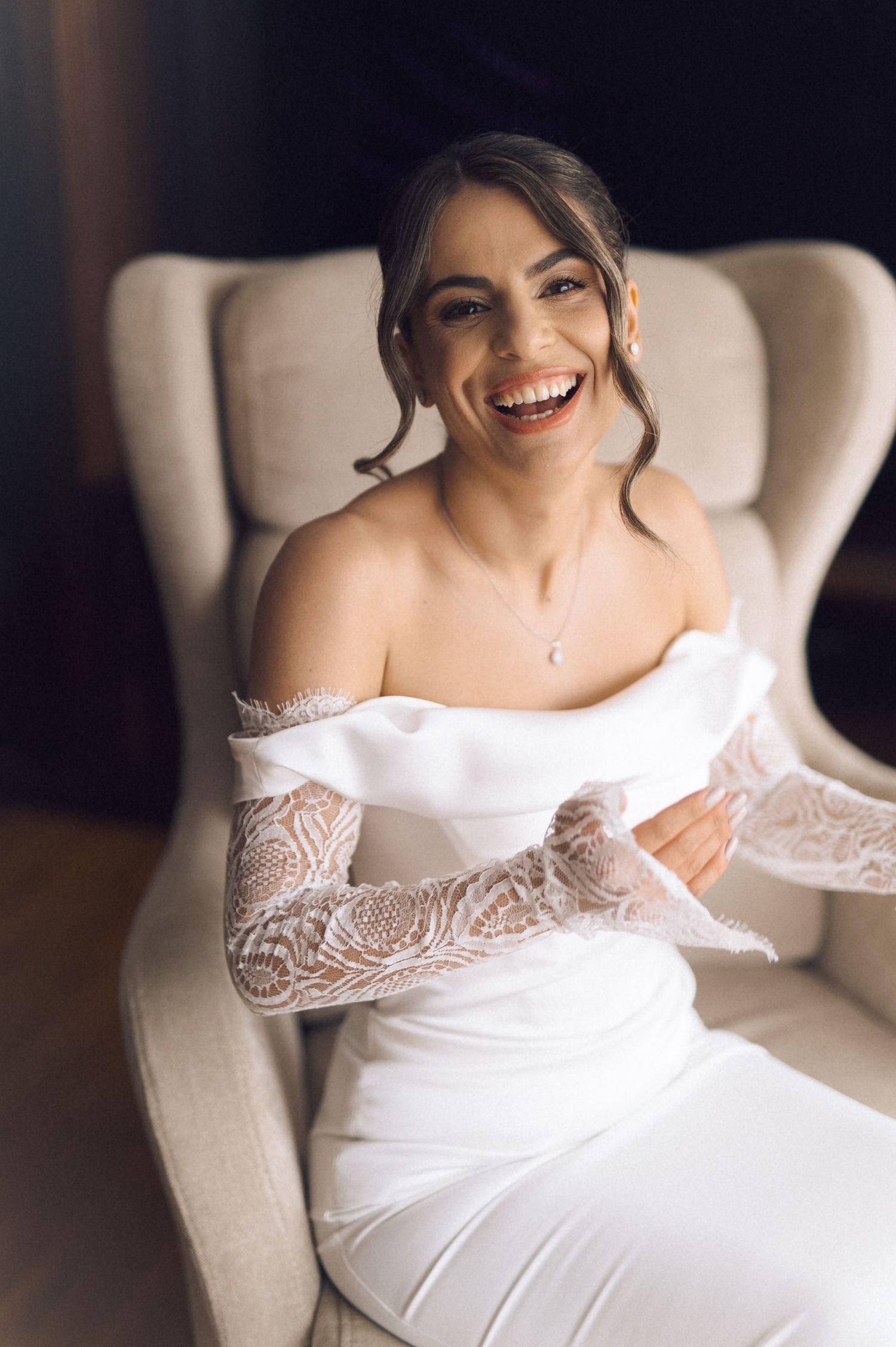 Smiling bride in off-shoulder white lace wedding gown seated in beige armchair, laughing joyfully on wedding day.