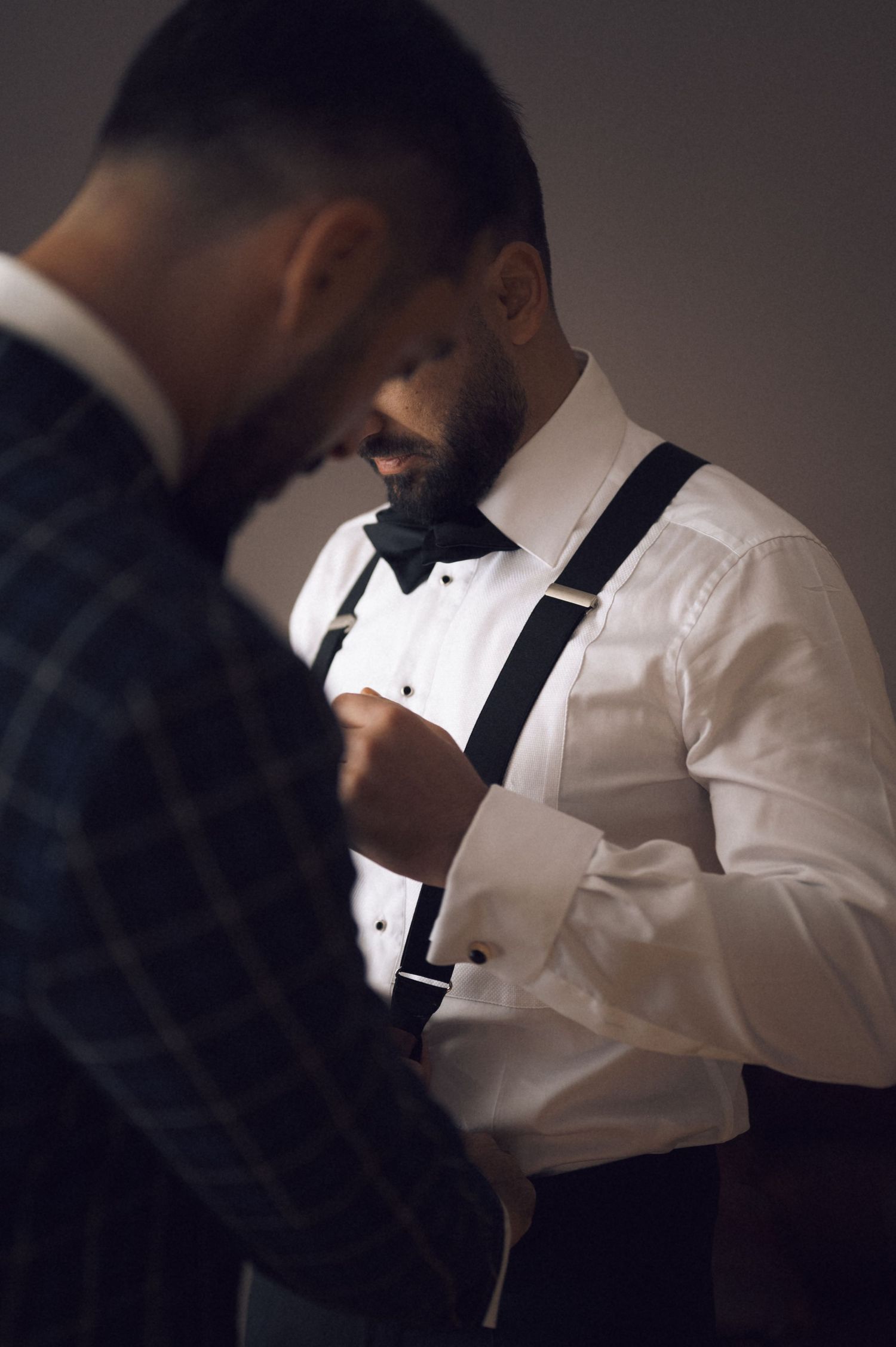 Bearded groom in white dress shirt with suspenders and bow tie fastening cufflinks with groomsman in plaid jacket nearby.