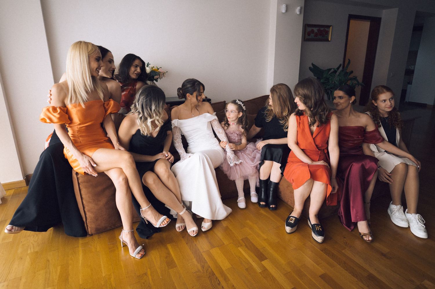 Bride in white gown surrounded by bridesmaids in colorful dresses sitting together on a sofa before the wedding.
