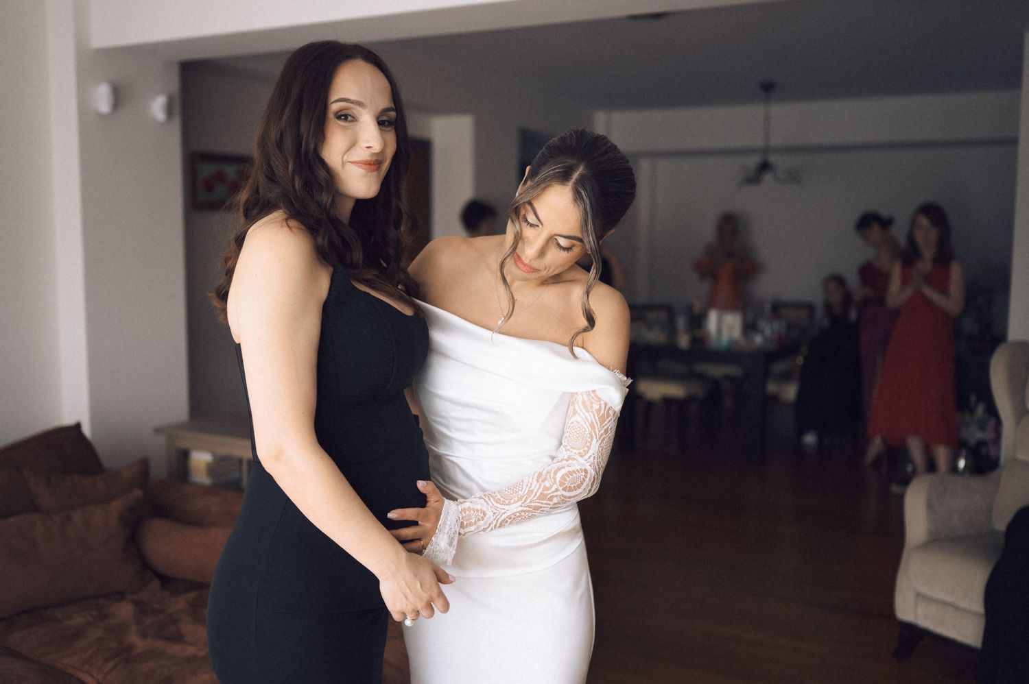 Bridesmaid in black dress helps bride in white off-shoulder gown prepare for wedding ceremony indoors.