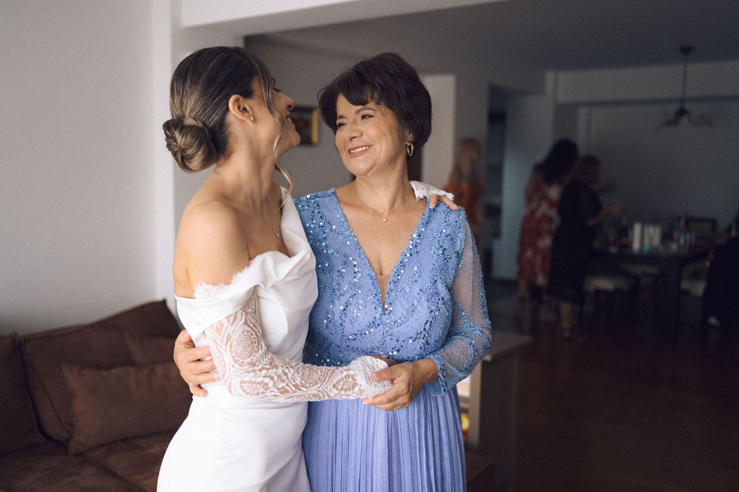 Bride in white lace gown embracing older woman in blue dress, sharing a joyful moment before wedding ceremony.