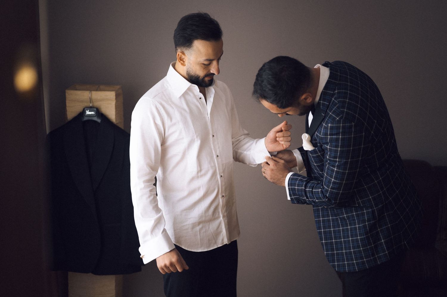 Man in white shirt gets cufflinks fastened by friend in plaid suit before formal event.