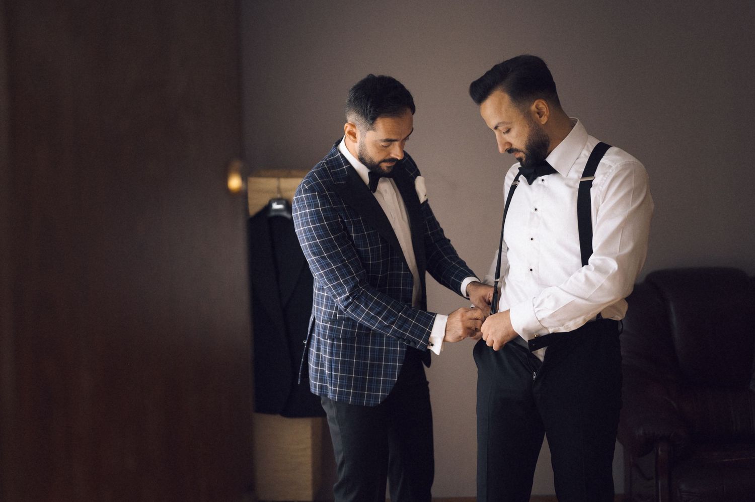 Two men in formal tuxedos help each other with cufflinks while getting ready for a wedding ceremony.