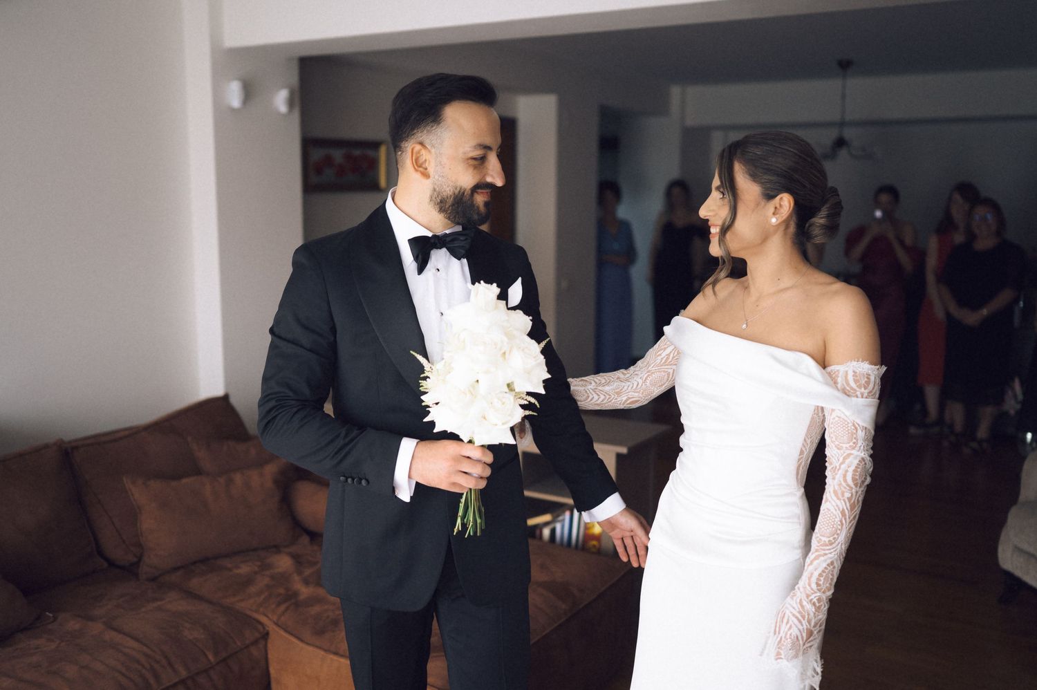 A groom in a black tuxedo holds white flowers while sharing a tender moment with his bride in a white lace gown.