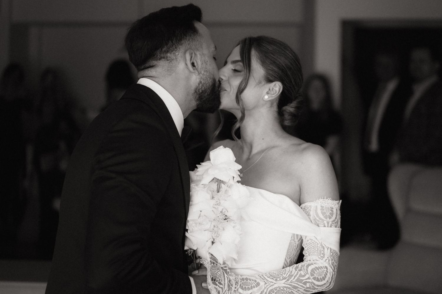 Black and white wedding photo of bride and groom sharing a kiss, bride in lace off-shoulder gown holding white flowers.