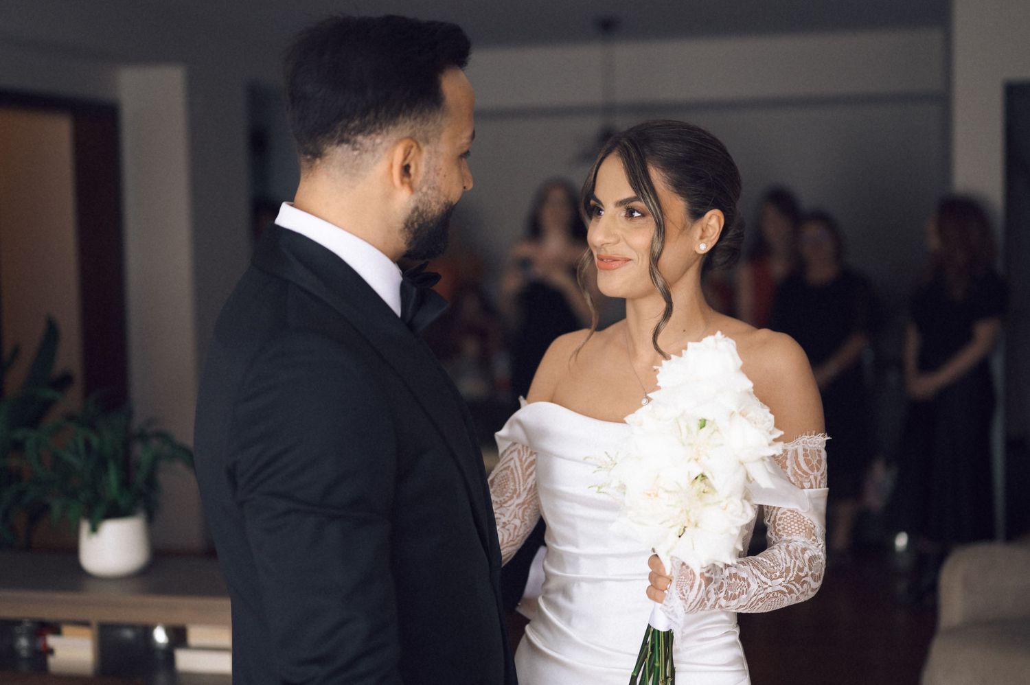 Bride in white off-shoulder gown holds white bouquet facing groom in black suit during indoor wedding ceremony.