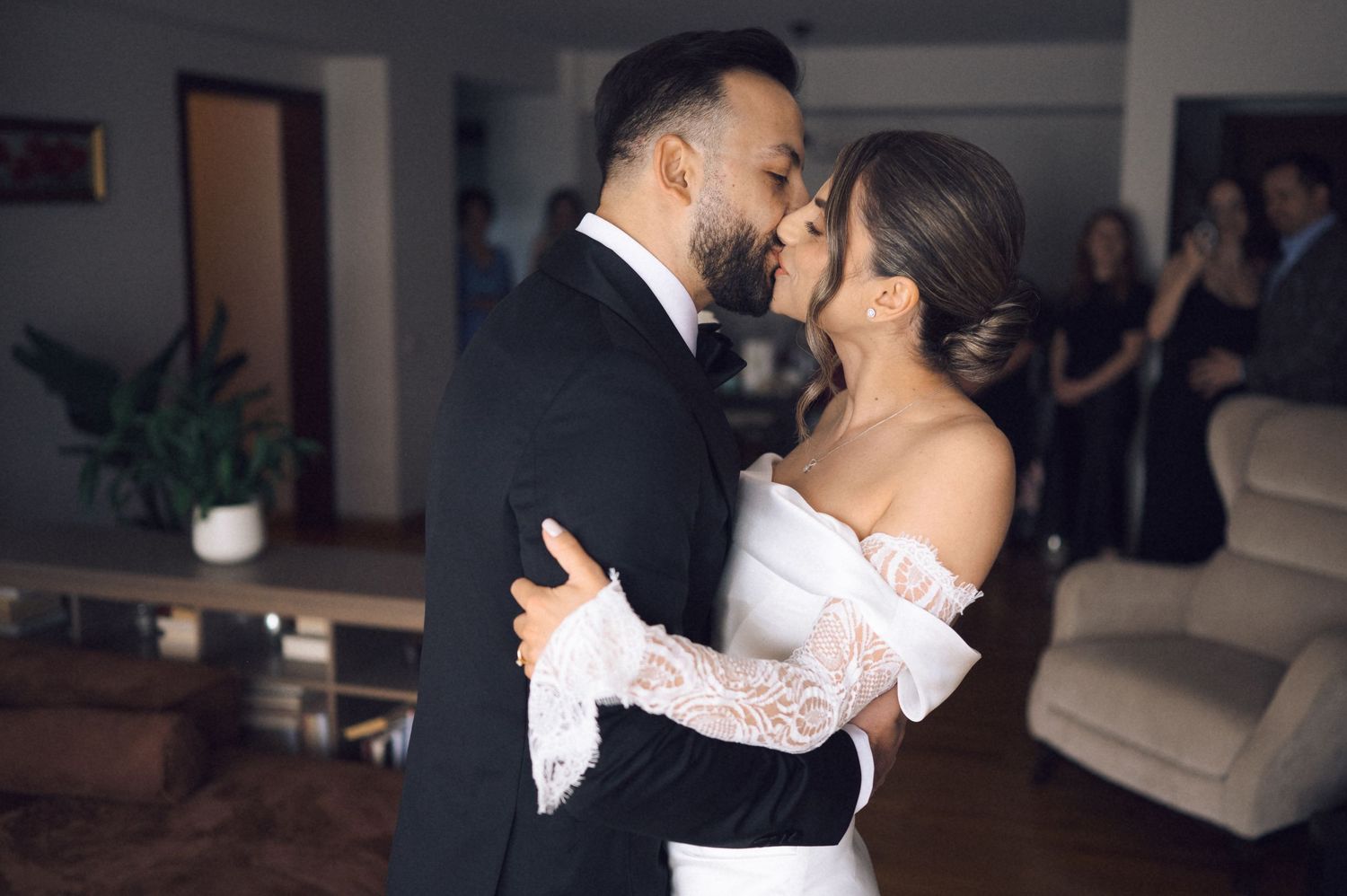 A couple shares a romantic first dance kiss at their wedding, bride in off-shoulder lace gown, guests watching in background.