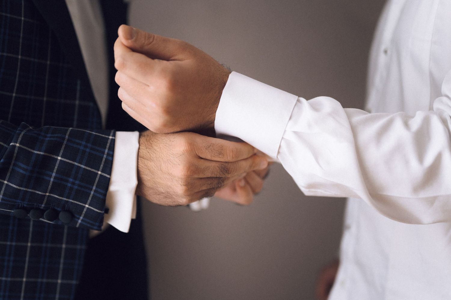 A person in a white shirt adjusts cufflinks on another mans suit jacket sleeve at a formal event.