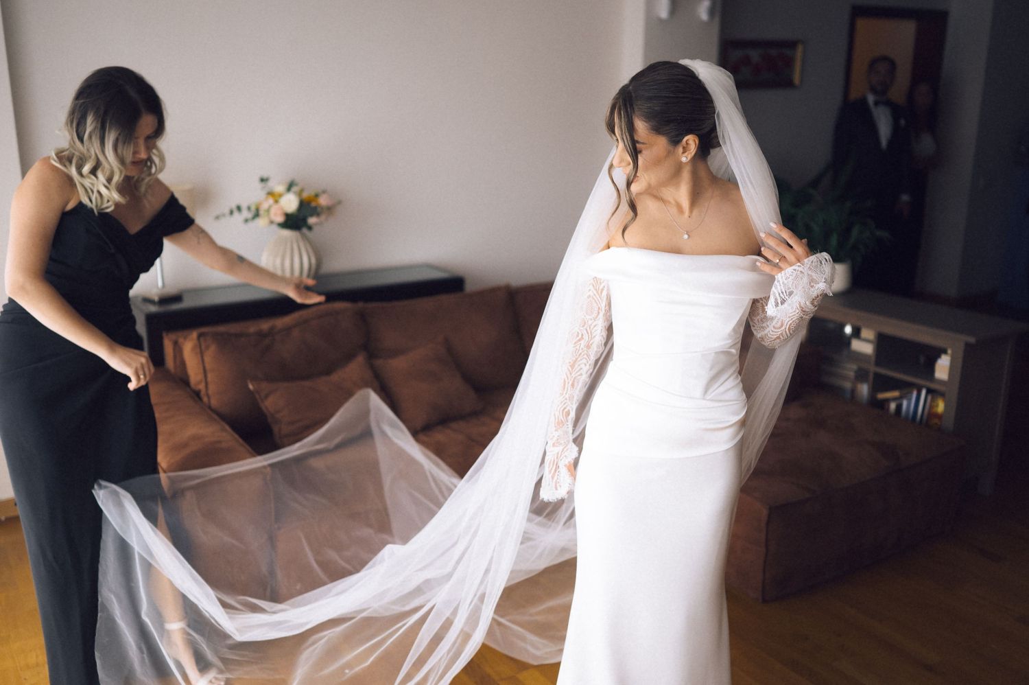 Bride in white off-shoulder gown with long veil being adjusted by bridesmaid in black dress indoors.