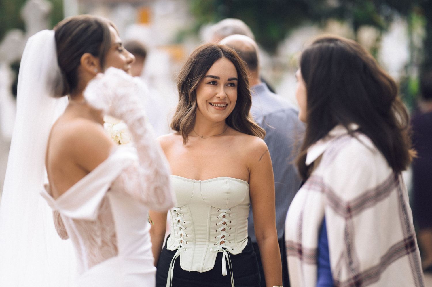 A young woman in a white corset top smiles outdoors near a bride in a wedding veil at a street event.
