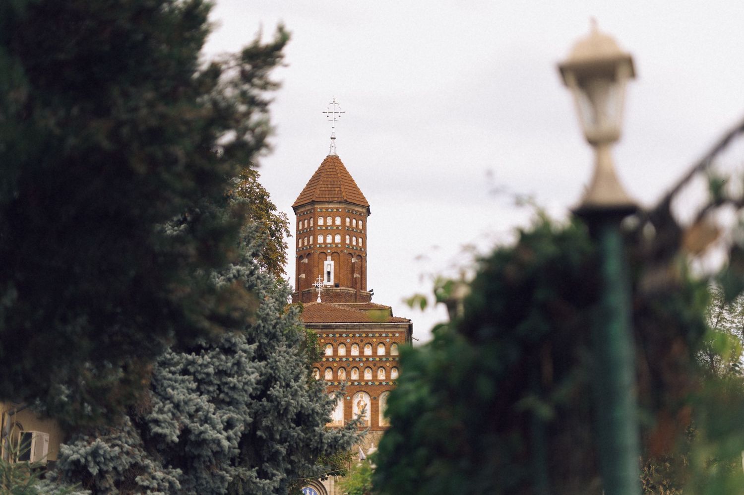 Historic brick church tower with a cross visible through lush green trees and a street lamp.