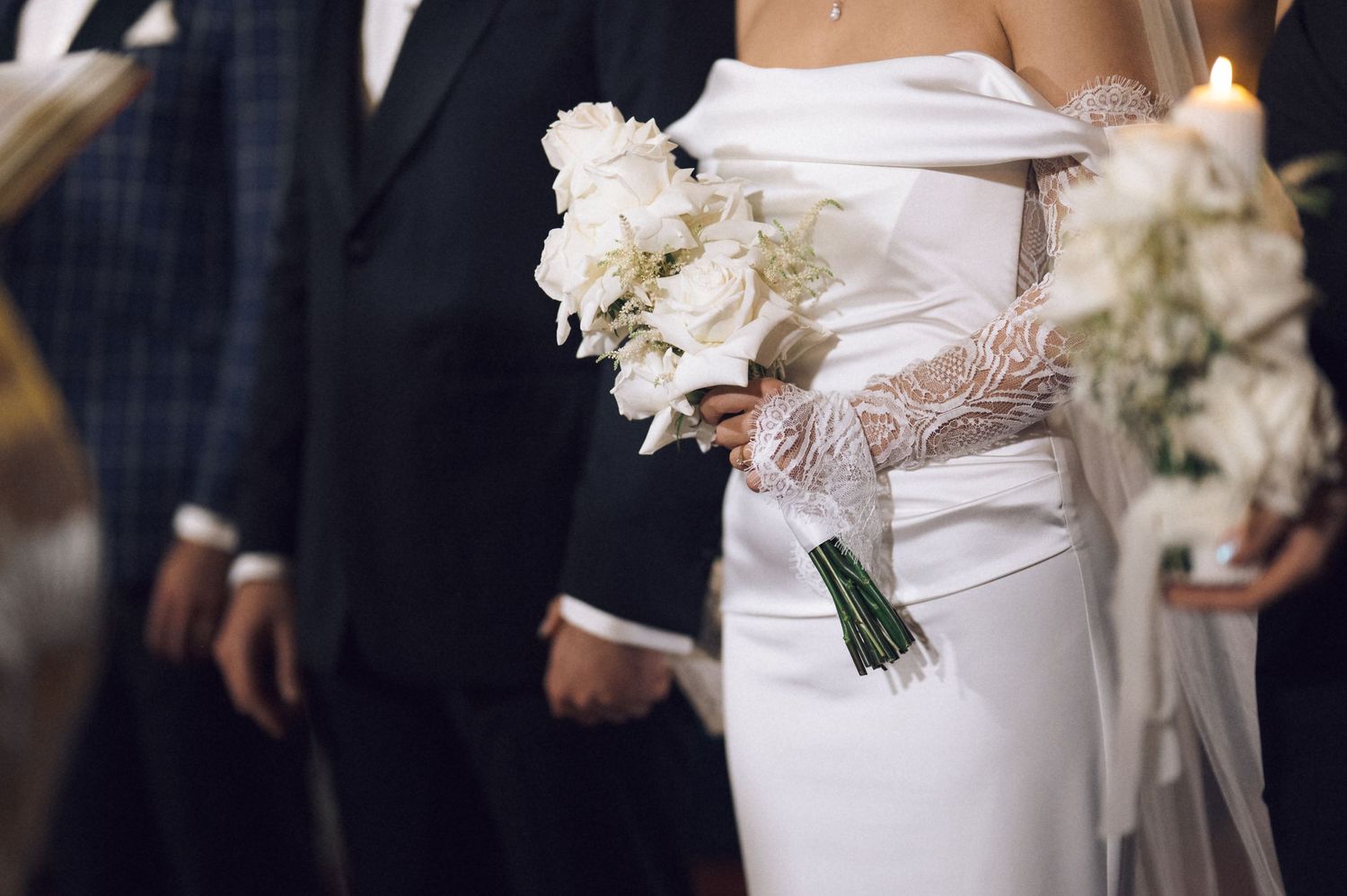 Bride in white off-shoulder gown holding elegant white rose bouquet stands beside groom in black suit at wedding ceremony.