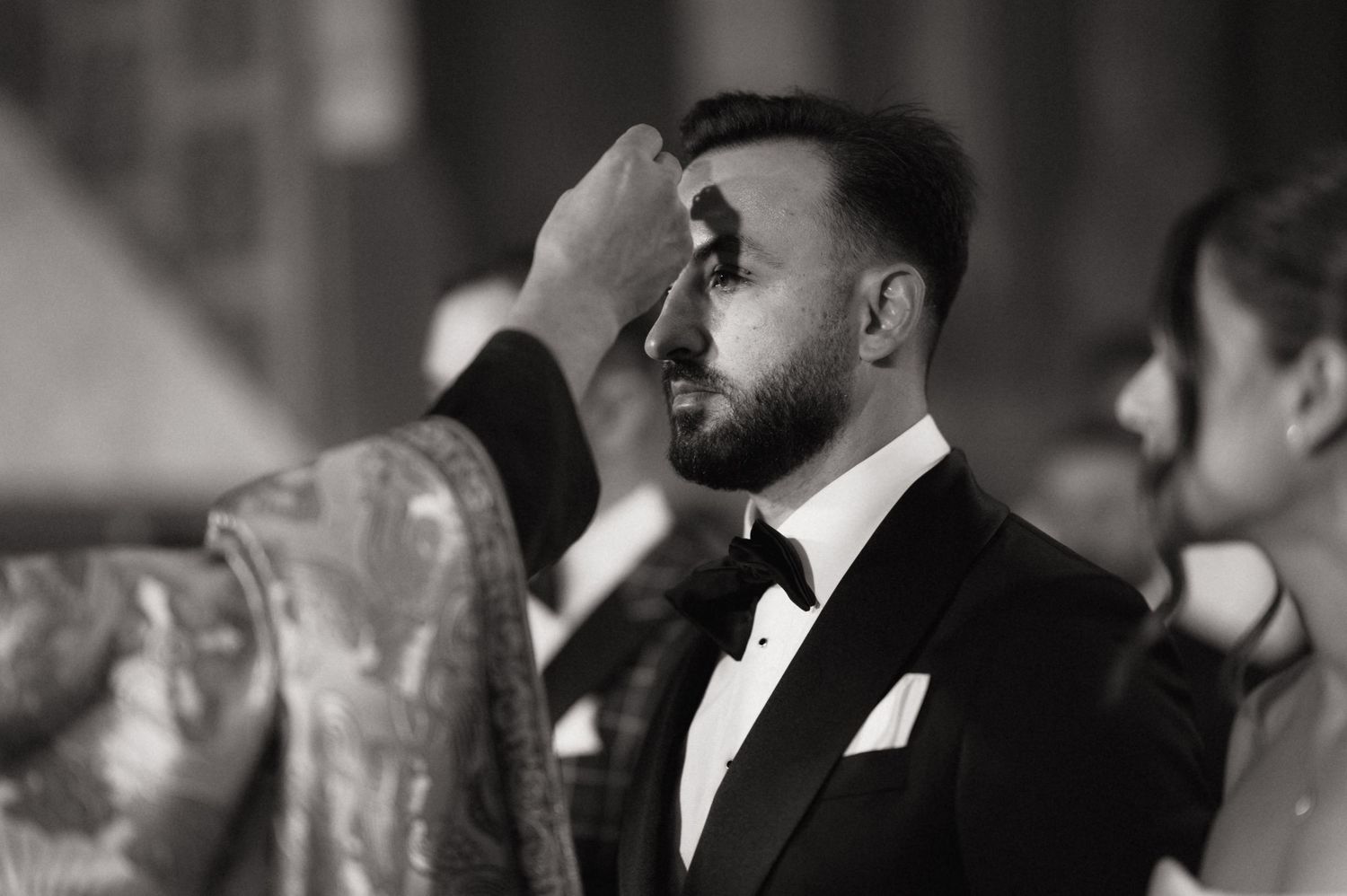 Black and white photo of a groom in tuxedo receiving a blessing during a wedding ceremony.