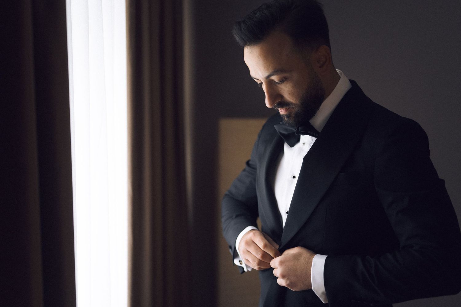A well-dressed man in a black tuxedo with bow tie adjusts his jacket near a window in soft natural light.
