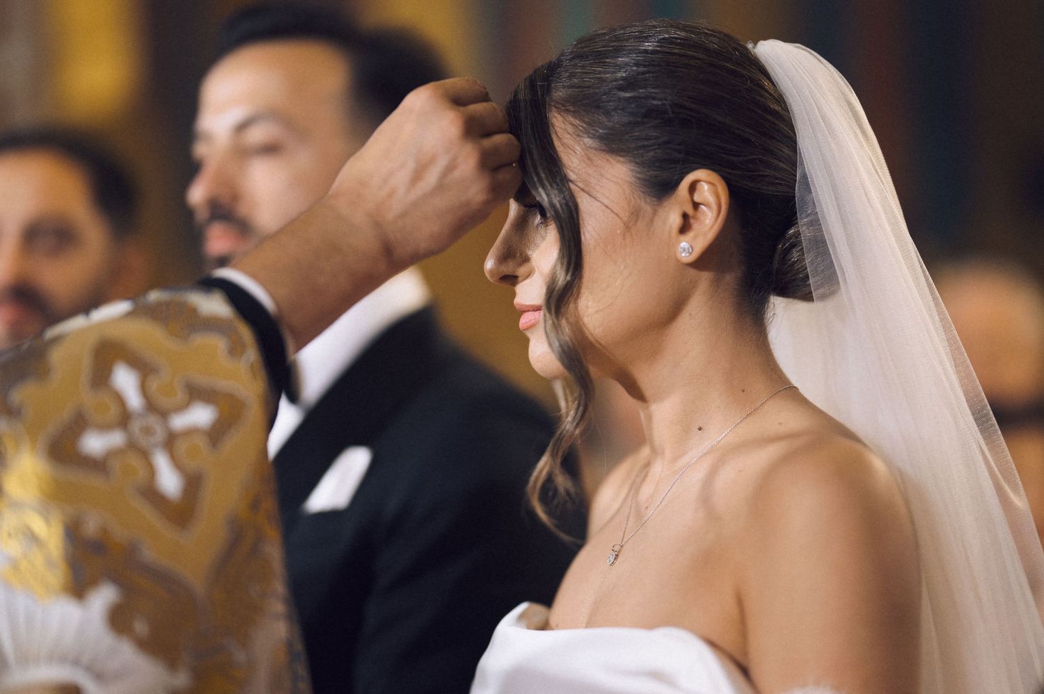 Bride in white veil receives blessing from officiant during elegant wedding ceremony.