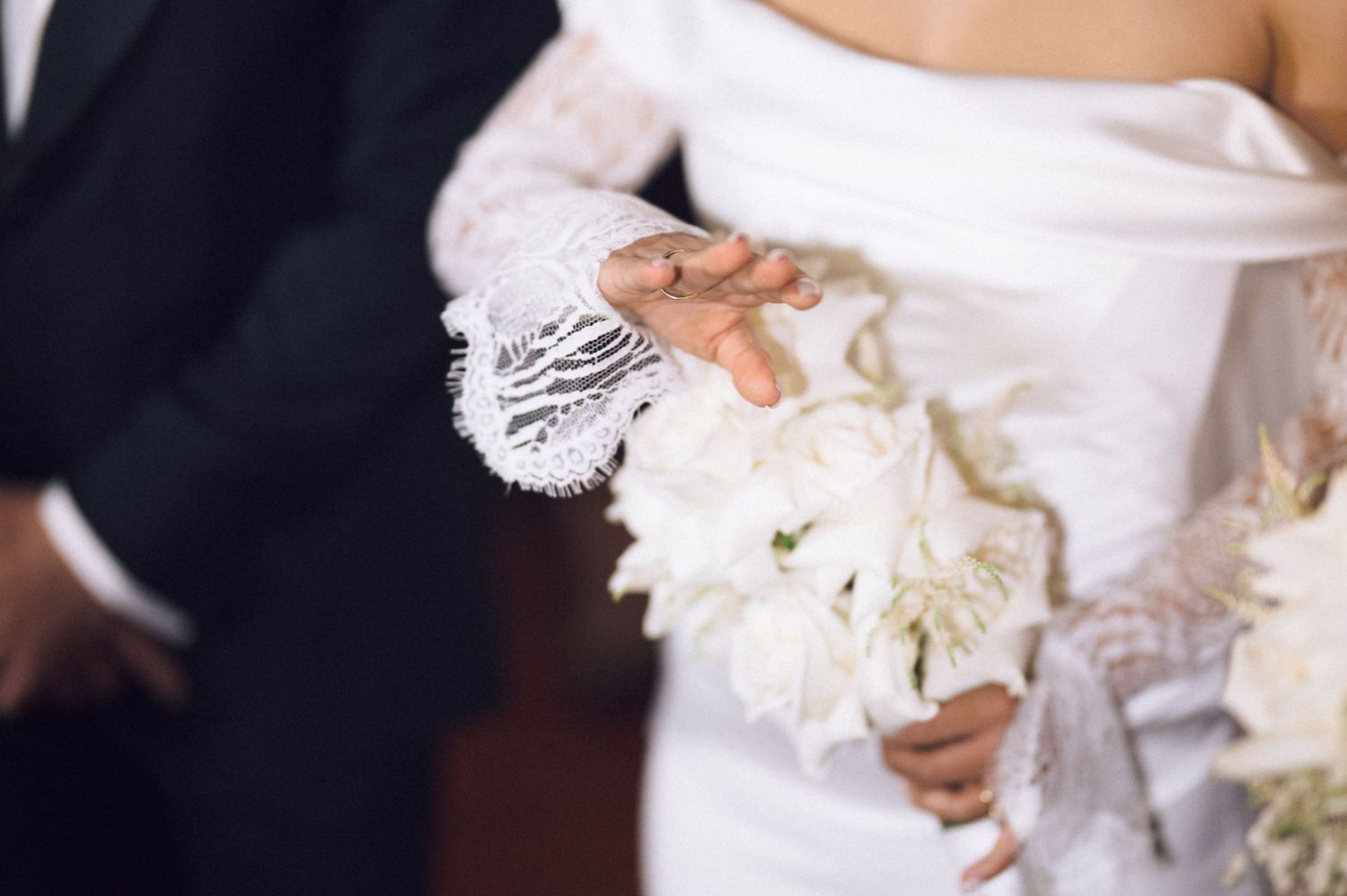 Bride in white lace gown holding elegant white floral bouquet beside groom in black suit at wedding ceremony.