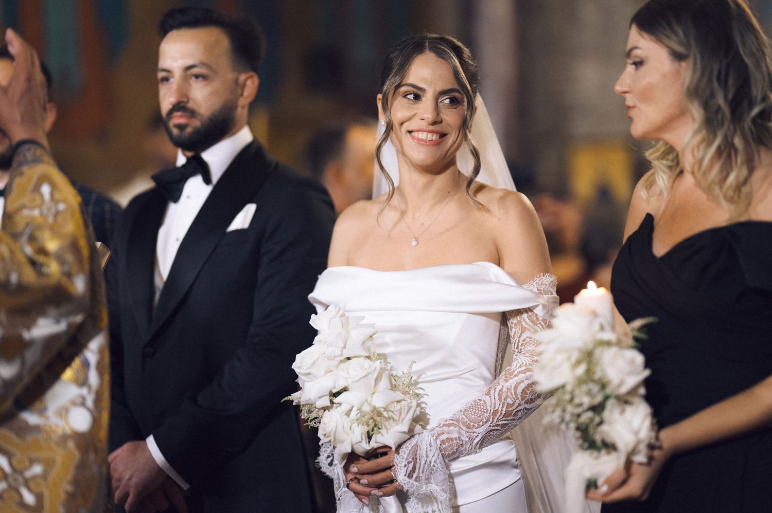 Bride in off-shoulder white gown holds bouquet at altar with groom in tuxedo and bridesmaid in black dress.