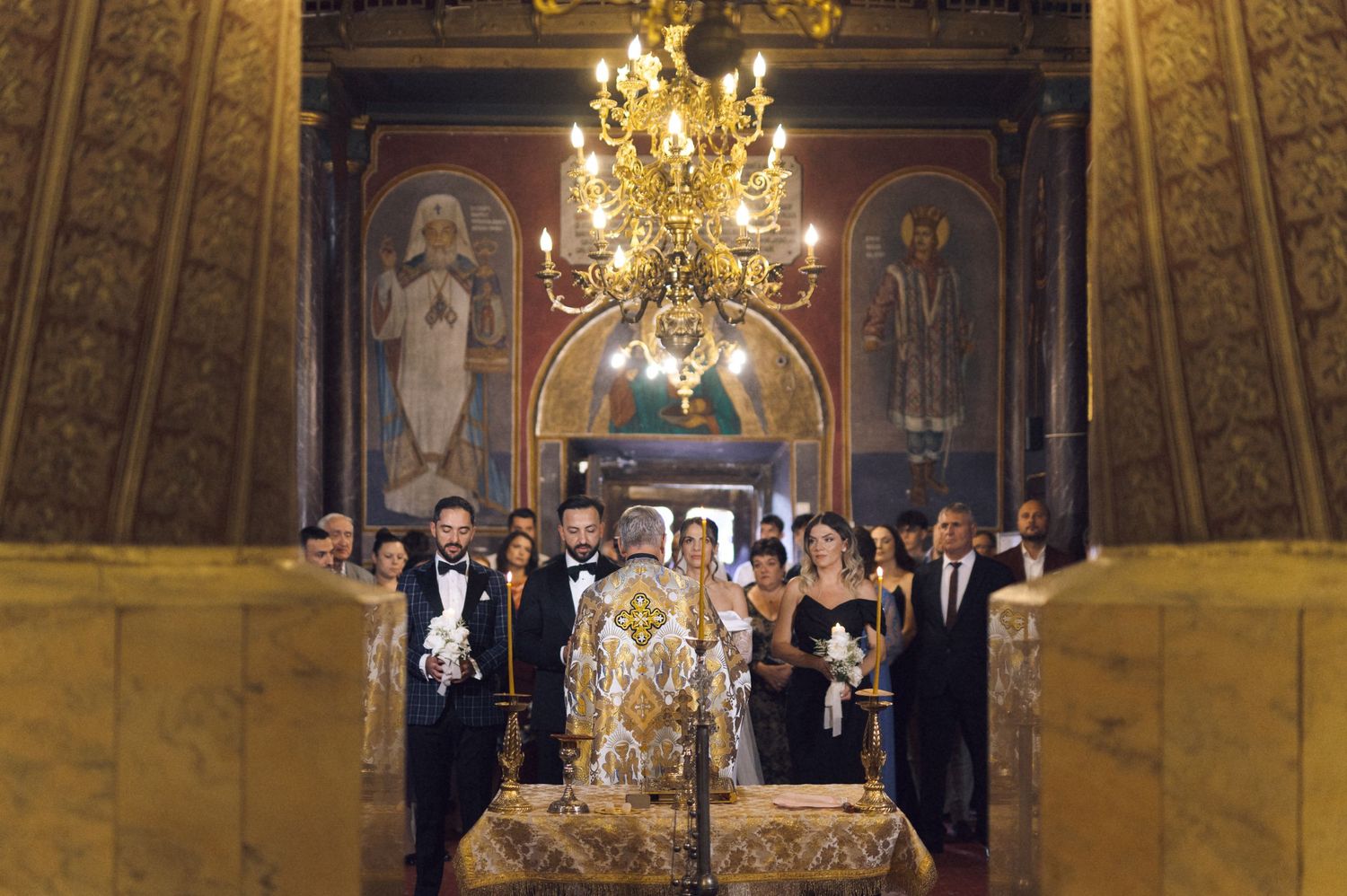 Orthodox church wedding ceremony with bridal party standing beneath golden chandelier amid Byzantine frescoes.