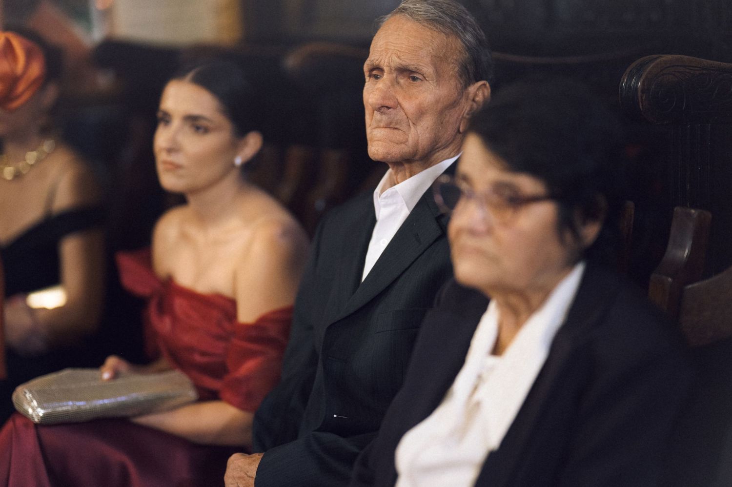 Three people seated in a row at a formal event, including an elderly man in a suit flanked by two women.