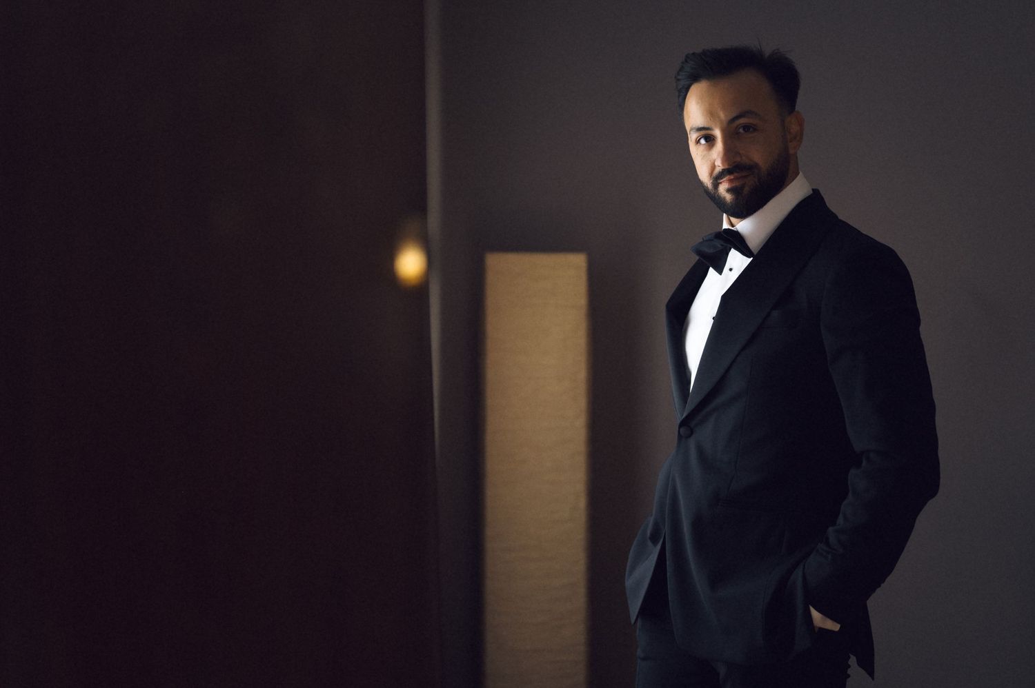 A sophisticated man in a black tuxedo with bow tie poses confidently in a dimly lit elegant setting.