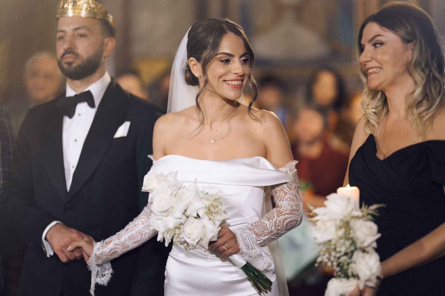 Bride in white off-shoulder gown with lace gloves smiles at wedding ceremony, flanked by groom and bridesmaid.