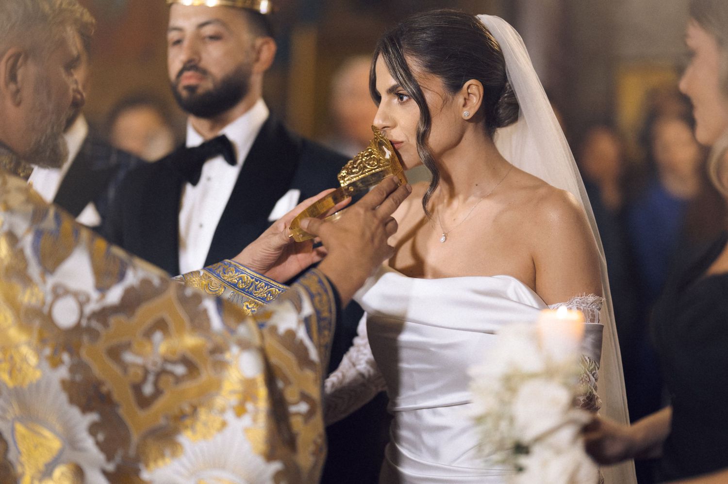 Bride receiving communion during an Orthodox wedding ceremony, wearing a white off-shoulder gown and veil.