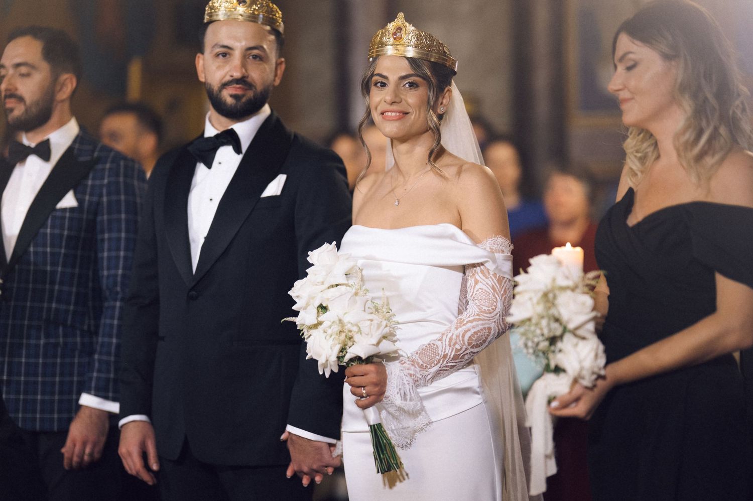 Bride and groom wearing gold crowns at elegant wedding ceremony, bride holding white bouquet in off-shoulder gown.