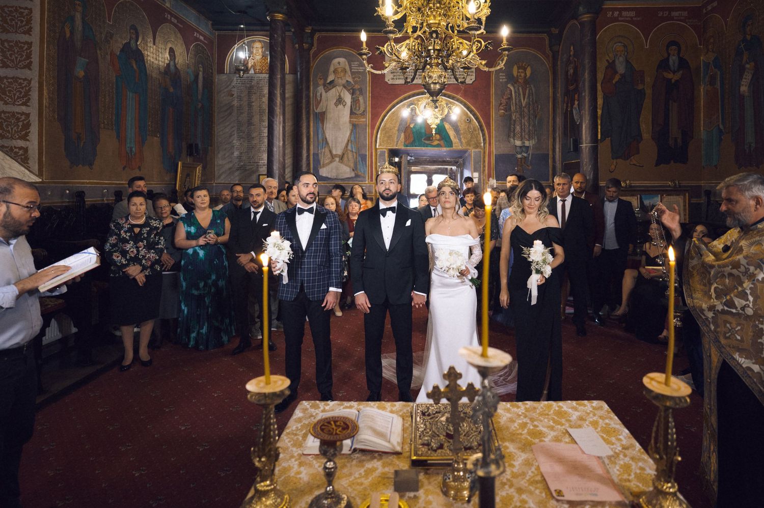 Bride and groom stand at the altar during an Orthodox church wedding ceremony surrounded by guests and golden candlesticks.