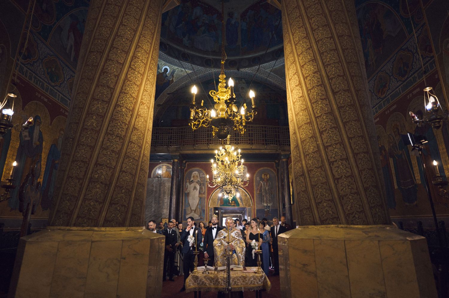 Orthodox ceremony inside ornate church with chandelier and clergy gathered around altar under decorated dome.