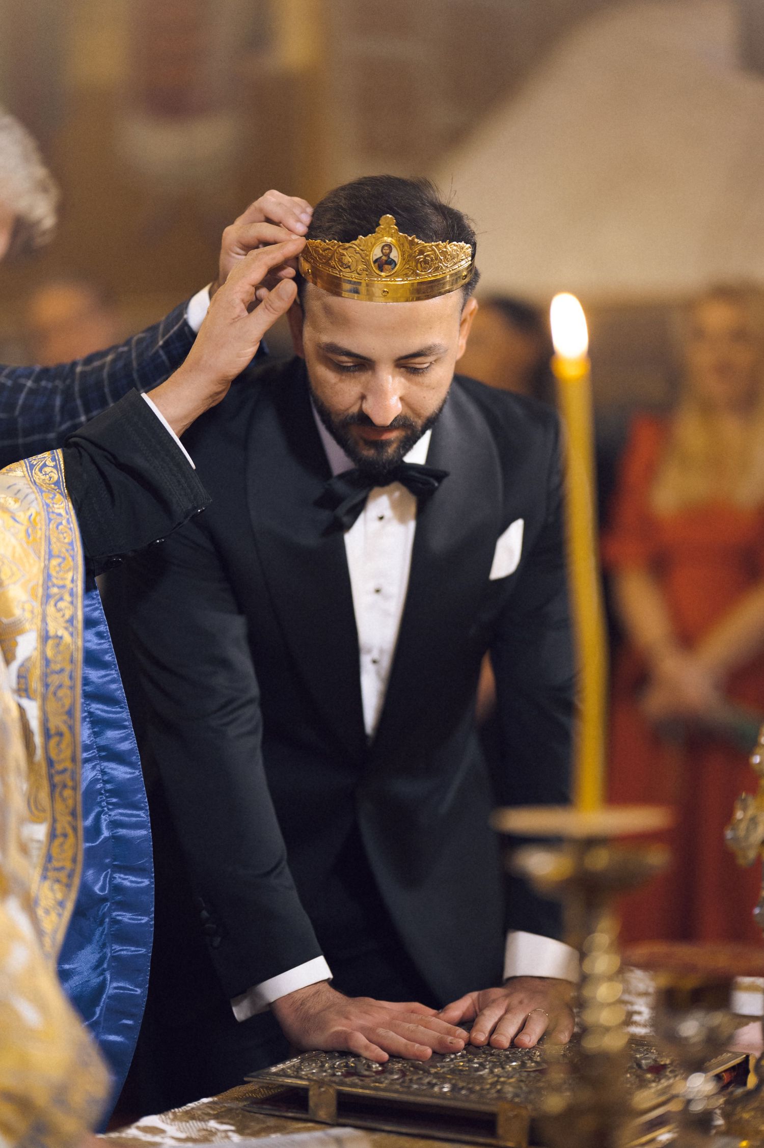 A man in a tuxedo receives a golden crown during an Orthodox wedding ceremony in a candlelit church.