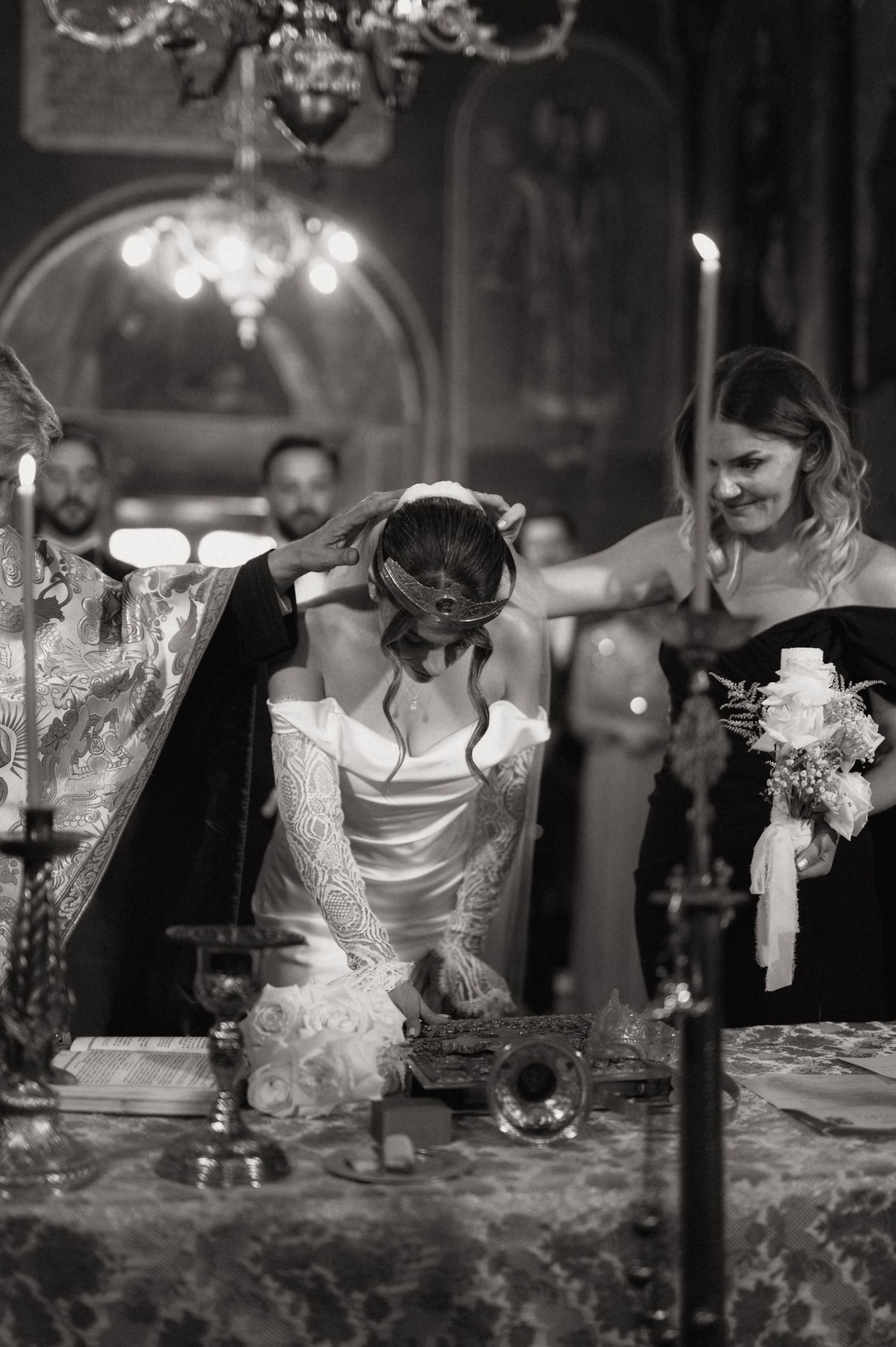 Black and white photo of a bride bowing during an Orthodox wedding ceremony with a smiling bridesmaid holding flowers nearby.