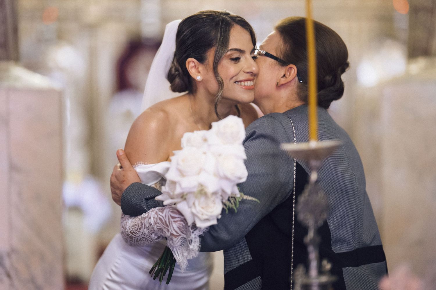 Bride in white gown hugging older woman at church wedding ceremony, holding bouquet of white roses.