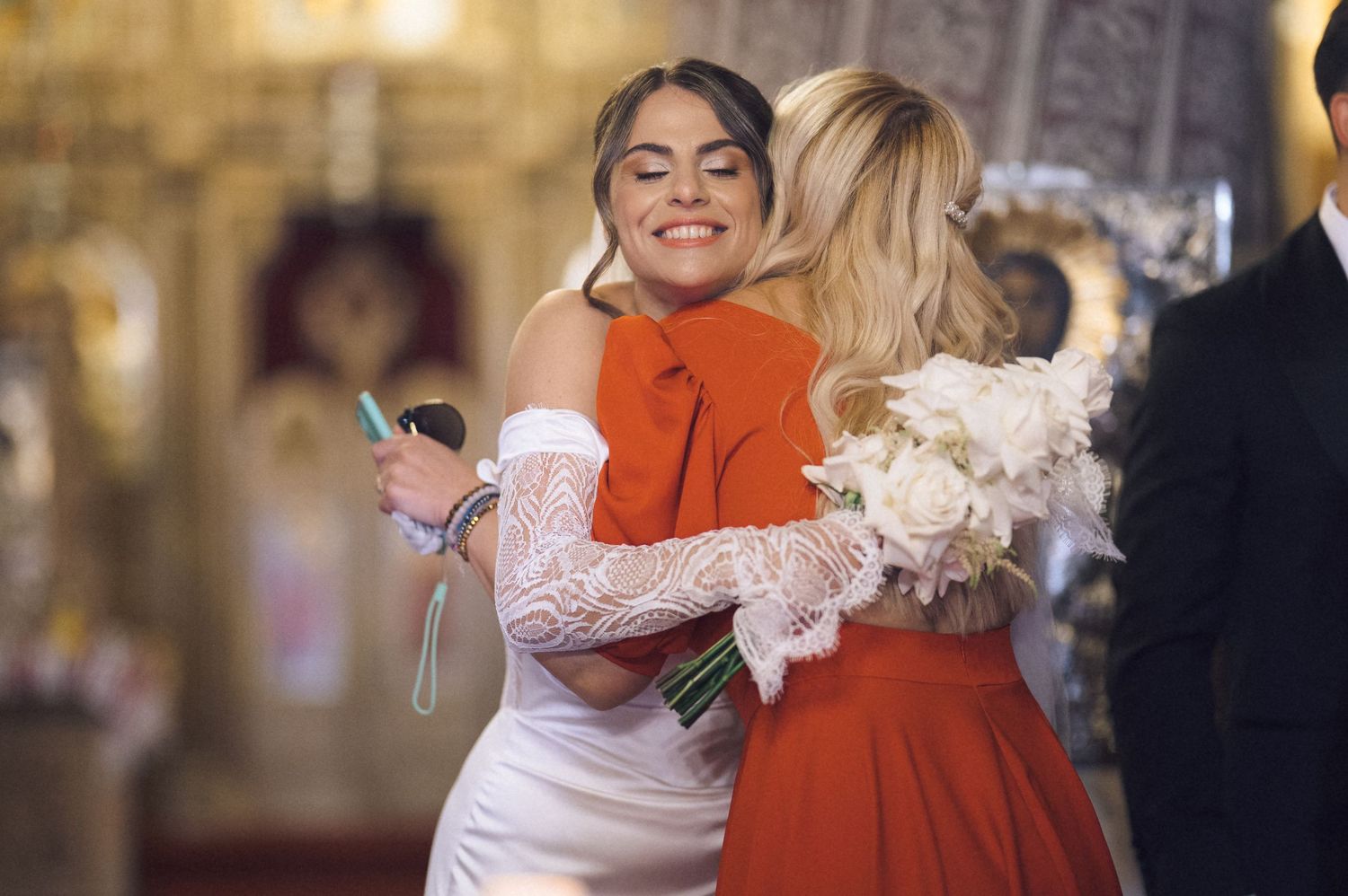 Bride in white lace dress embracing woman in red coat during church wedding celebration.
