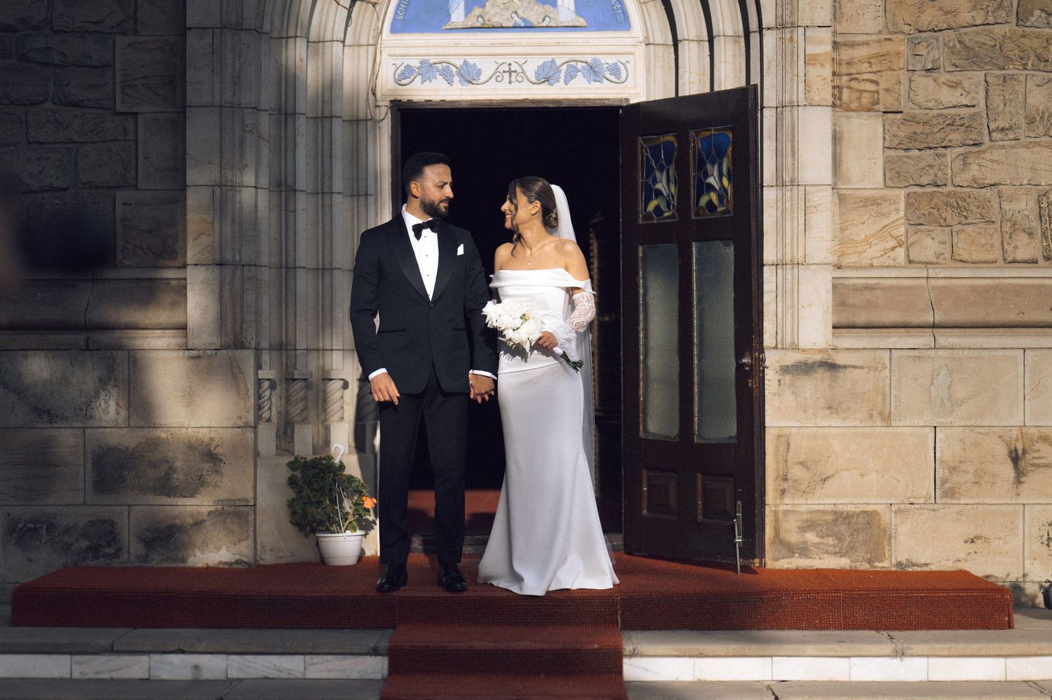 Bride and groom smiling at each other on church steps, she in white off-shoulder gown, he in black tuxedo.