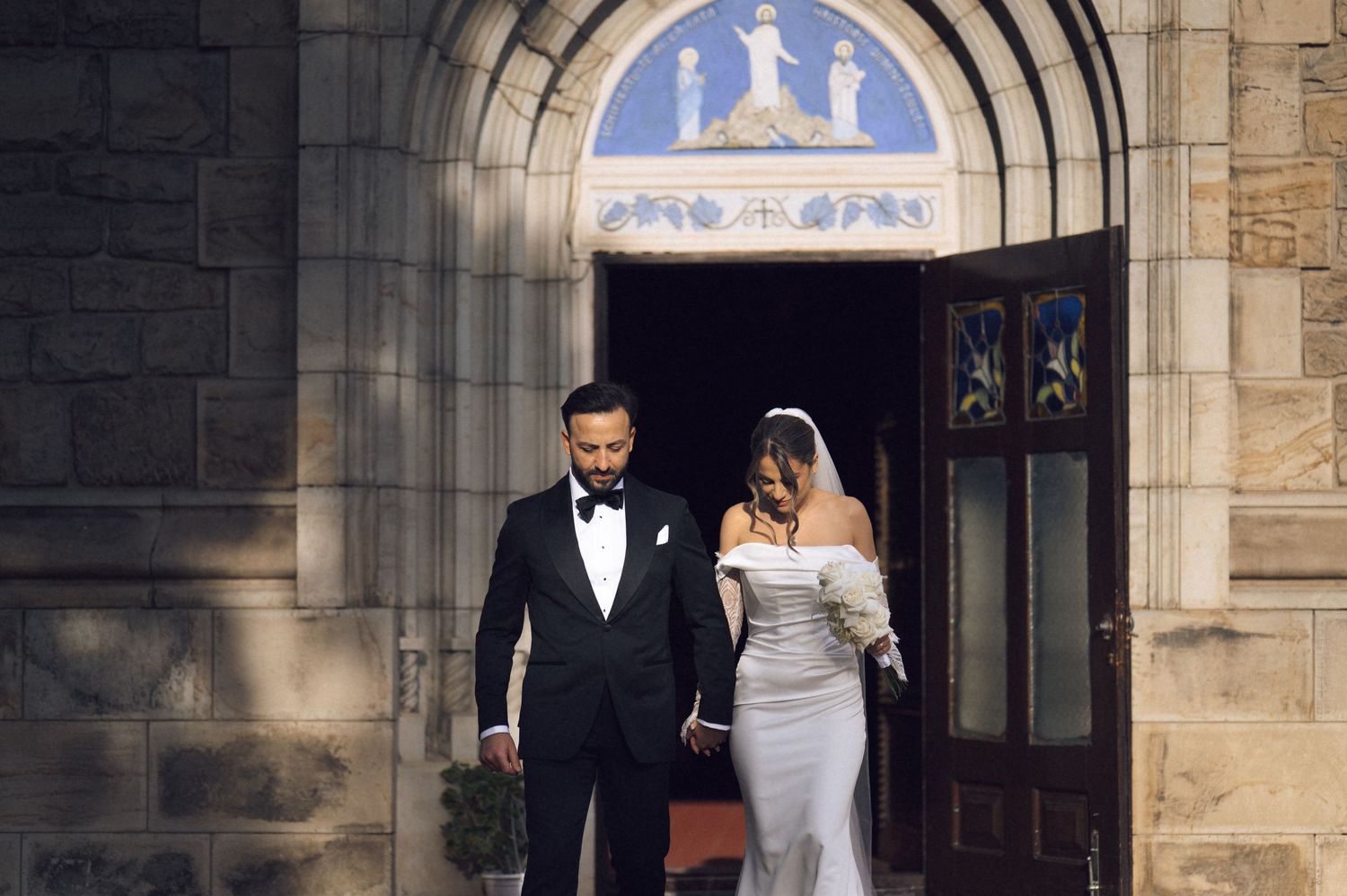 Elegant couple exiting stone church, bride in fitted white gown with veil, groom in classic black tuxedo.